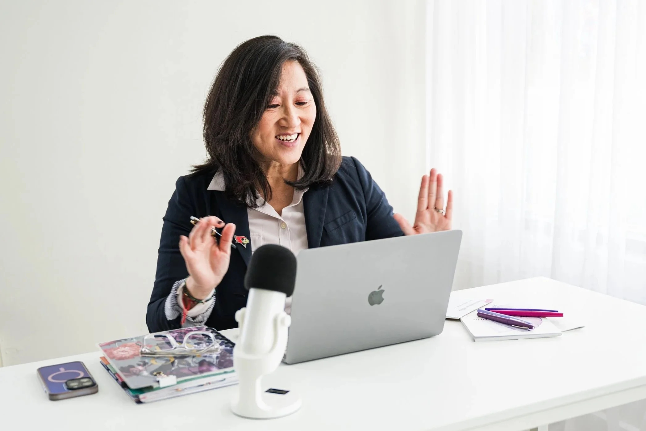 A woman smiling and waving at her laptop with a microphone in front of her, on a white desk with notebooks, pens, and glasses, in a bright room with sheer white curtains.