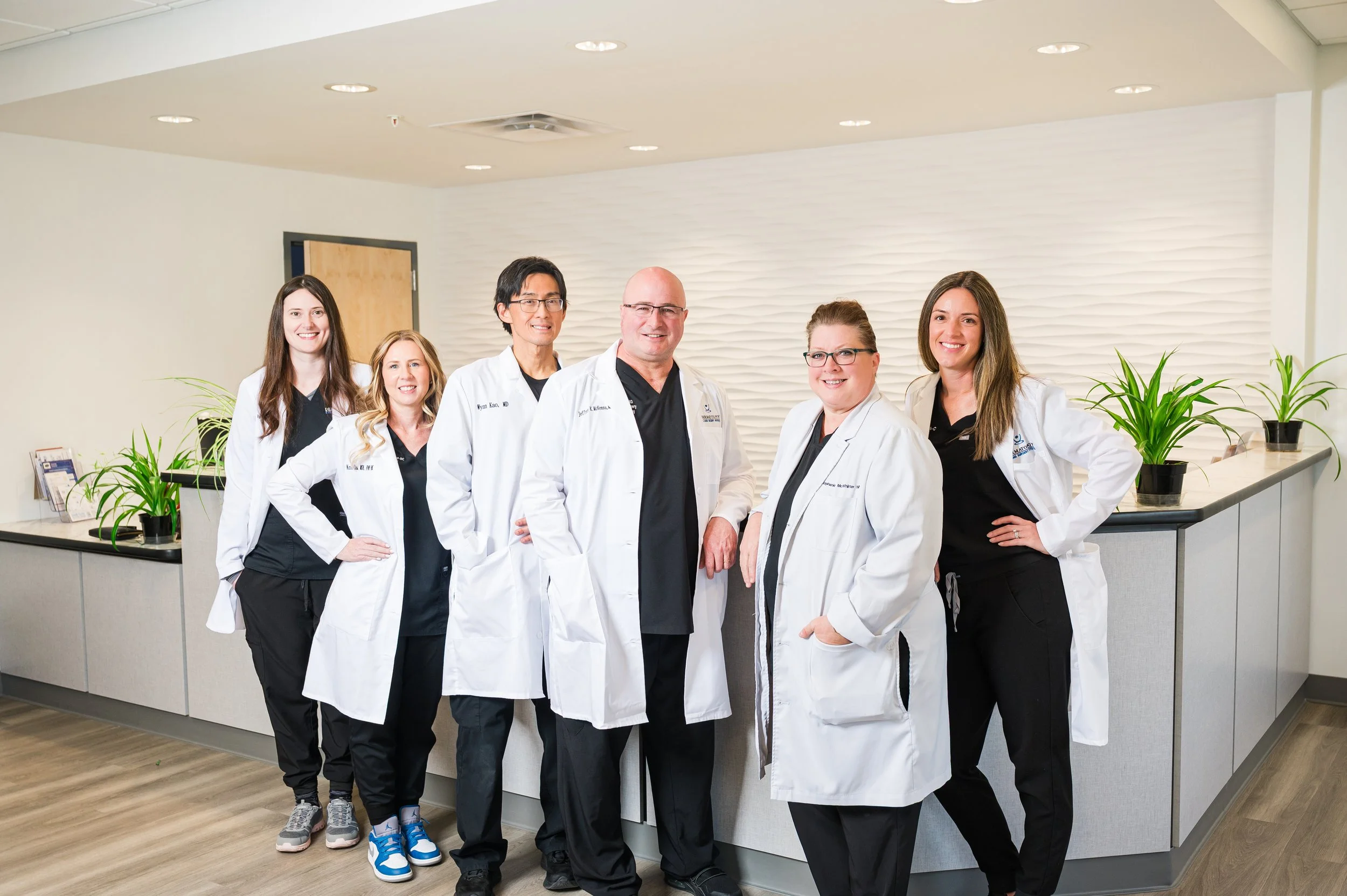 A group of seven healthcare professionals, including doctors and nurses, standing together in a modern medical facility lobby with a white textured wall, plants, and reception desk.