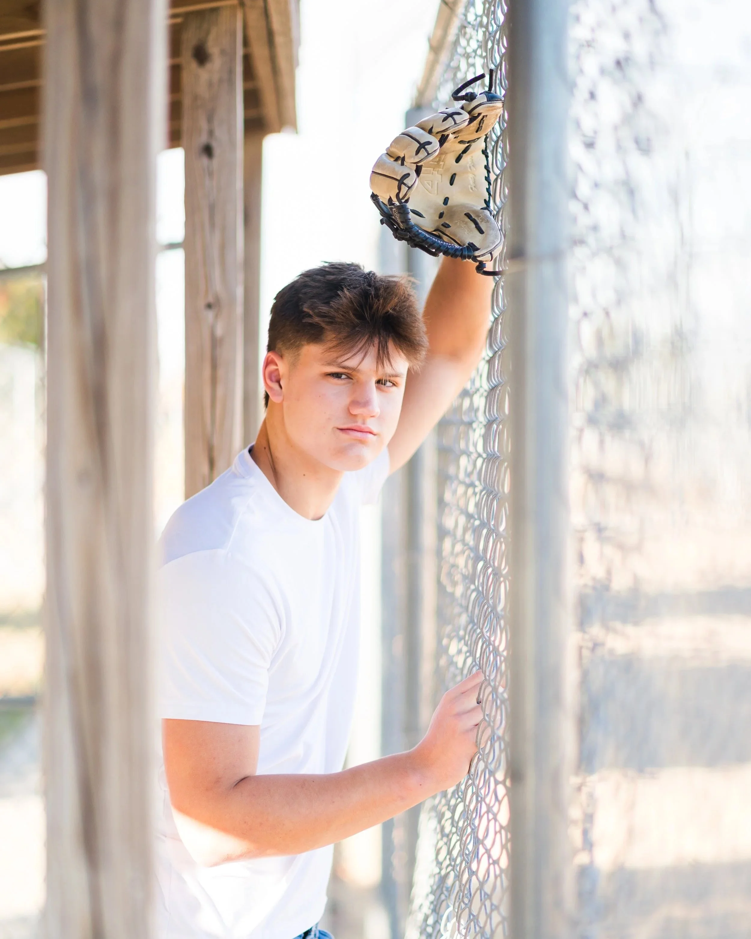 Young man standing next to a chain-link fence with a baseball glove resting on his hand, looking serious.