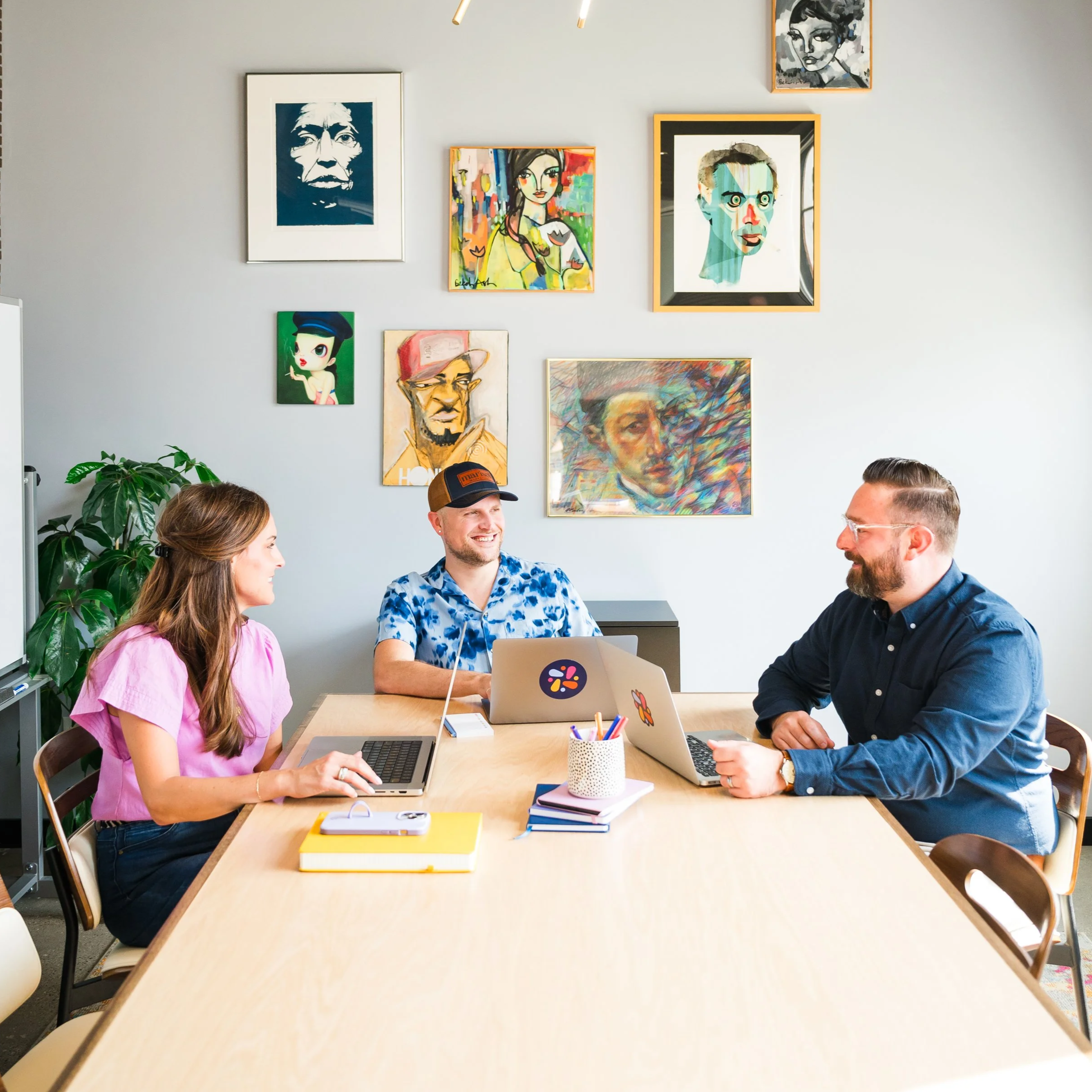 Three people sitting at a conference table in a room with colorful artwork on the wall, smiling and talking, with laptops and notebooks on the table.