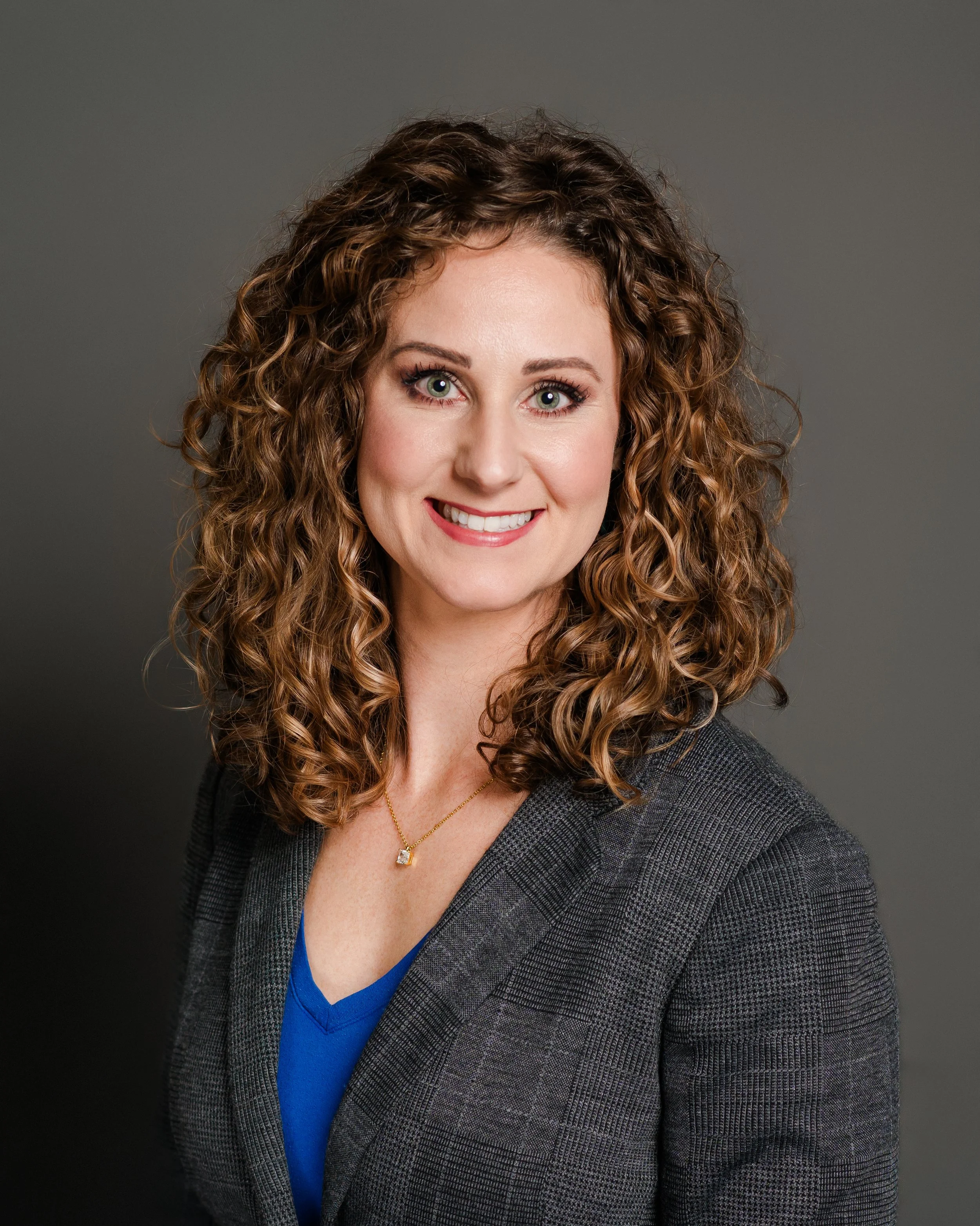 Headshot of a woman with curly brown hair, wearing a gray blazer over a blue top and a gold necklace, smiling against a gray background.