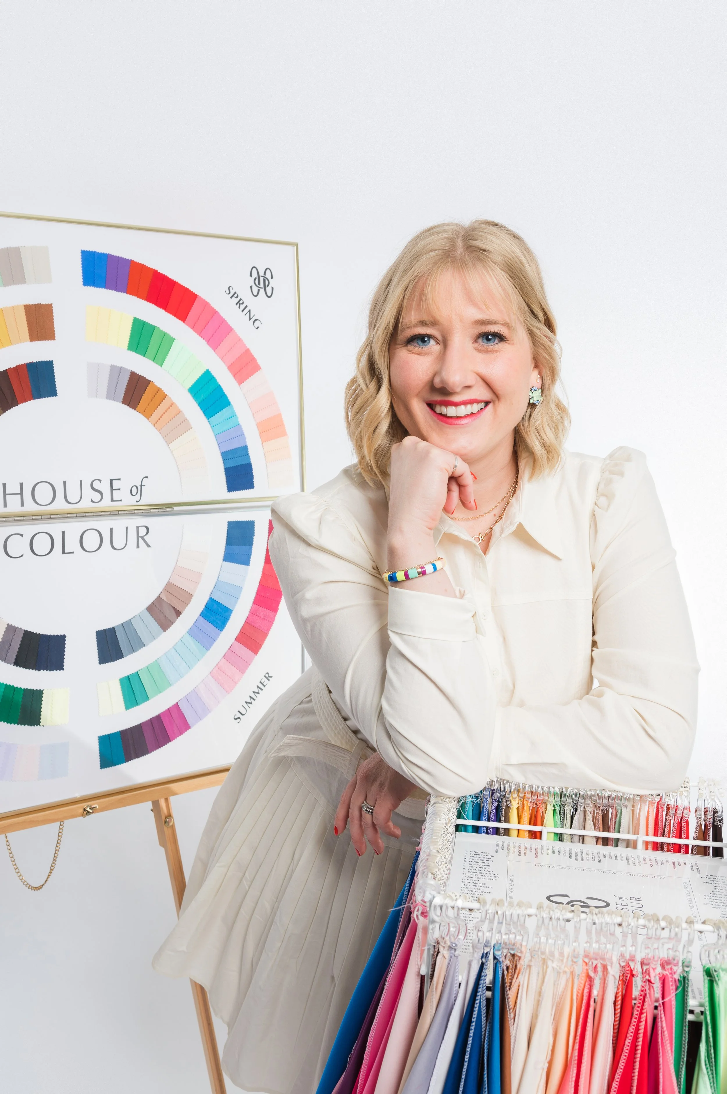 A woman with blonde hair smiling at the camera, sitting at a table with color swatches and fabric samples in front of her, in a room with a color wheel poster in the background.