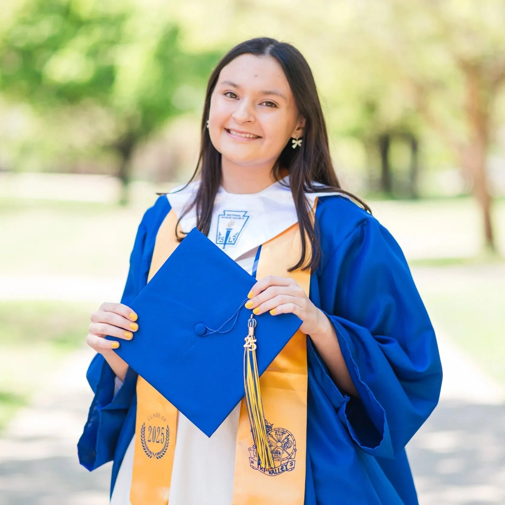 A young woman wearing a blue graduation gown and cap, holding her diploma, standing outdoors with blurred green trees in the background.