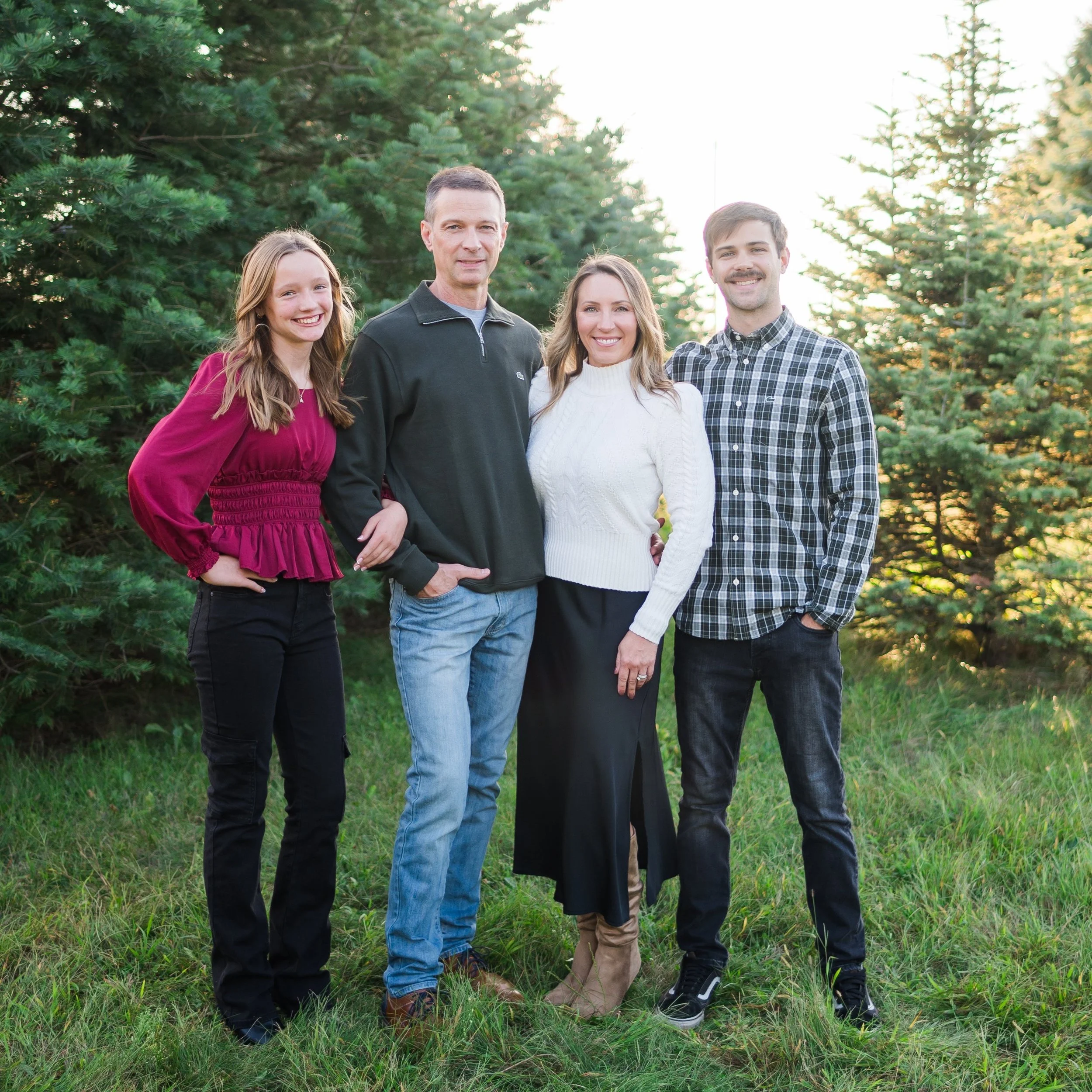 Family of four standing together outdoors in front of evergreen trees, smiling at the camera during sunset.