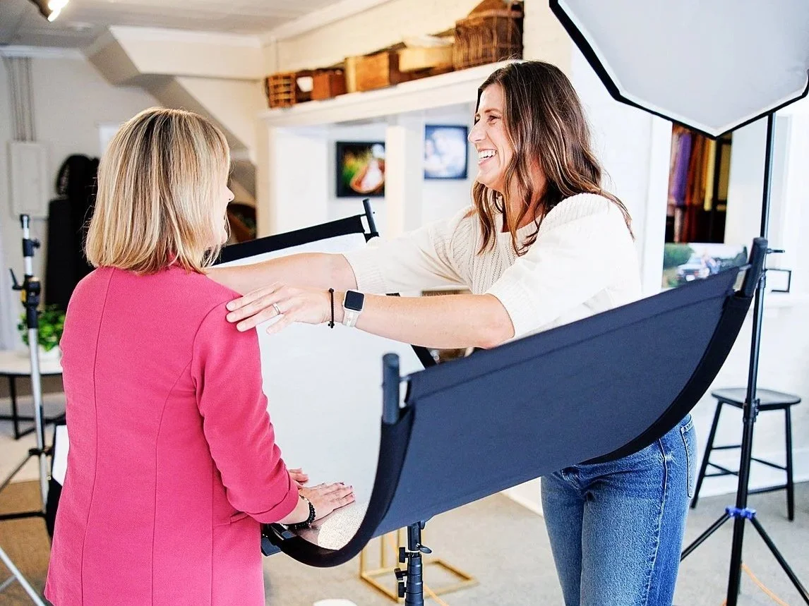 Two women engaging in a friendly conversation during a photo shoot, with professional lighting equipment in a spacious indoor studio.