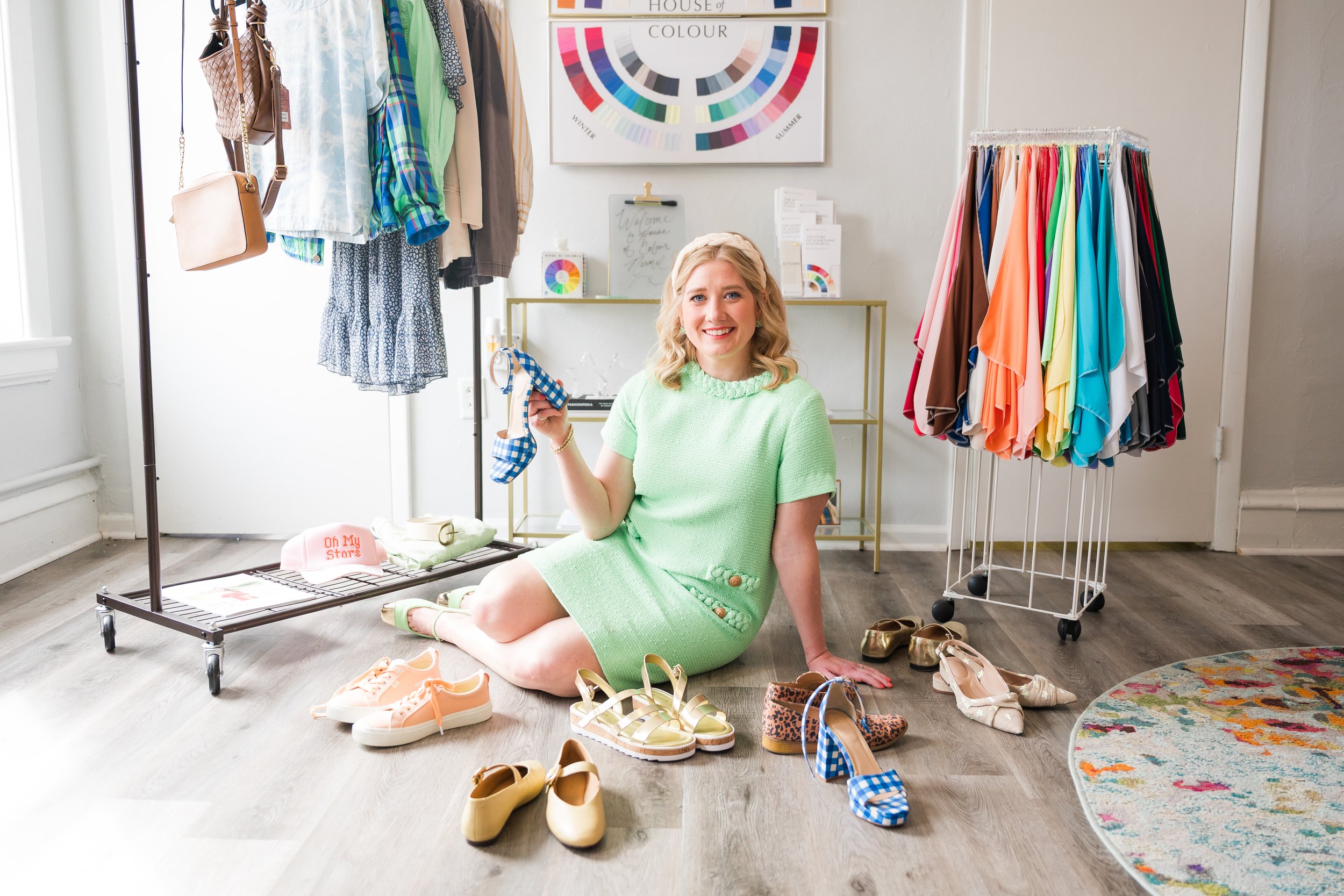 A young woman sitting on the floor of a boutique or clothing store, holding a blue and white checkered shoe, surrounded by various shoes and clothing racks with colorful garments. She is smiling and wearing a light green dress.