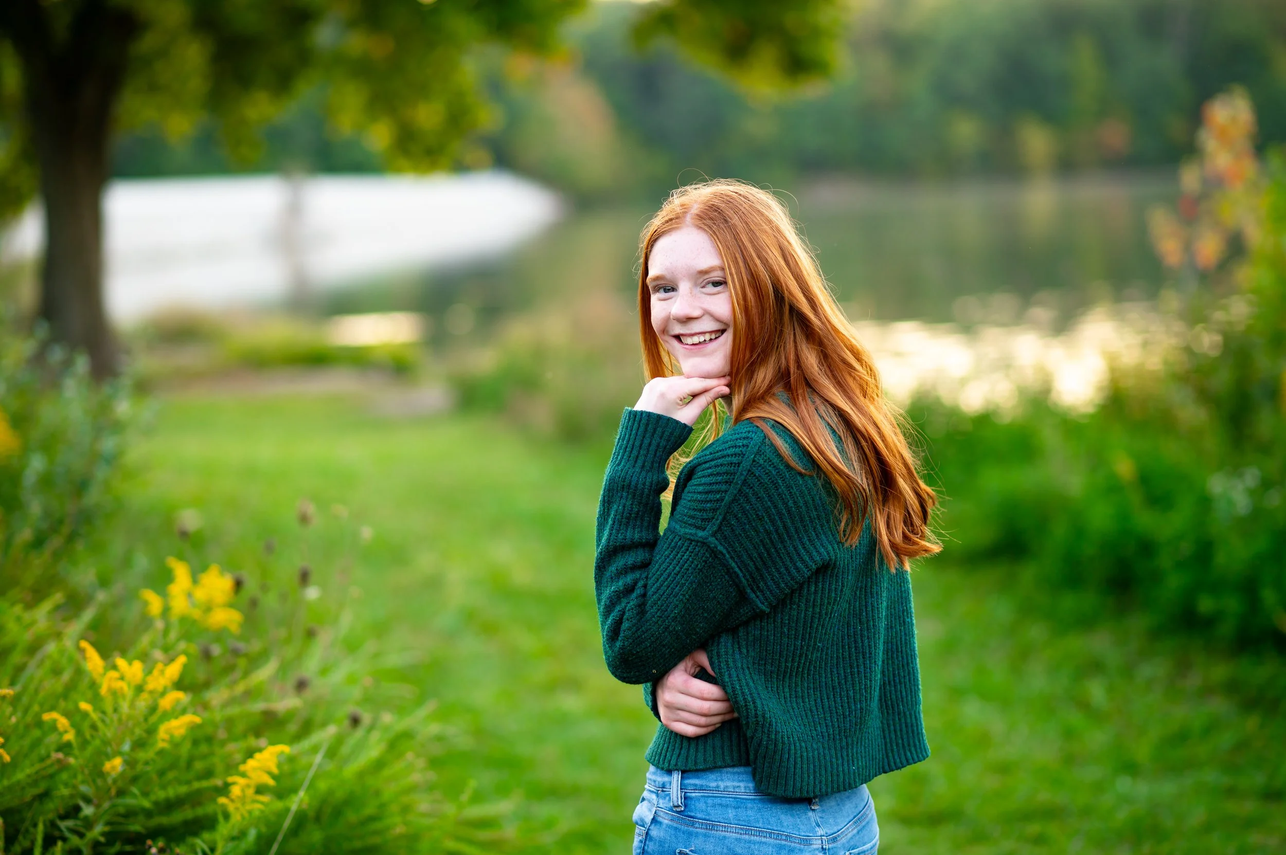 Young woman with red hair smiling outdoors near a lake, wearing a green sweater and jeans, with trees and yellow flowers in the background.