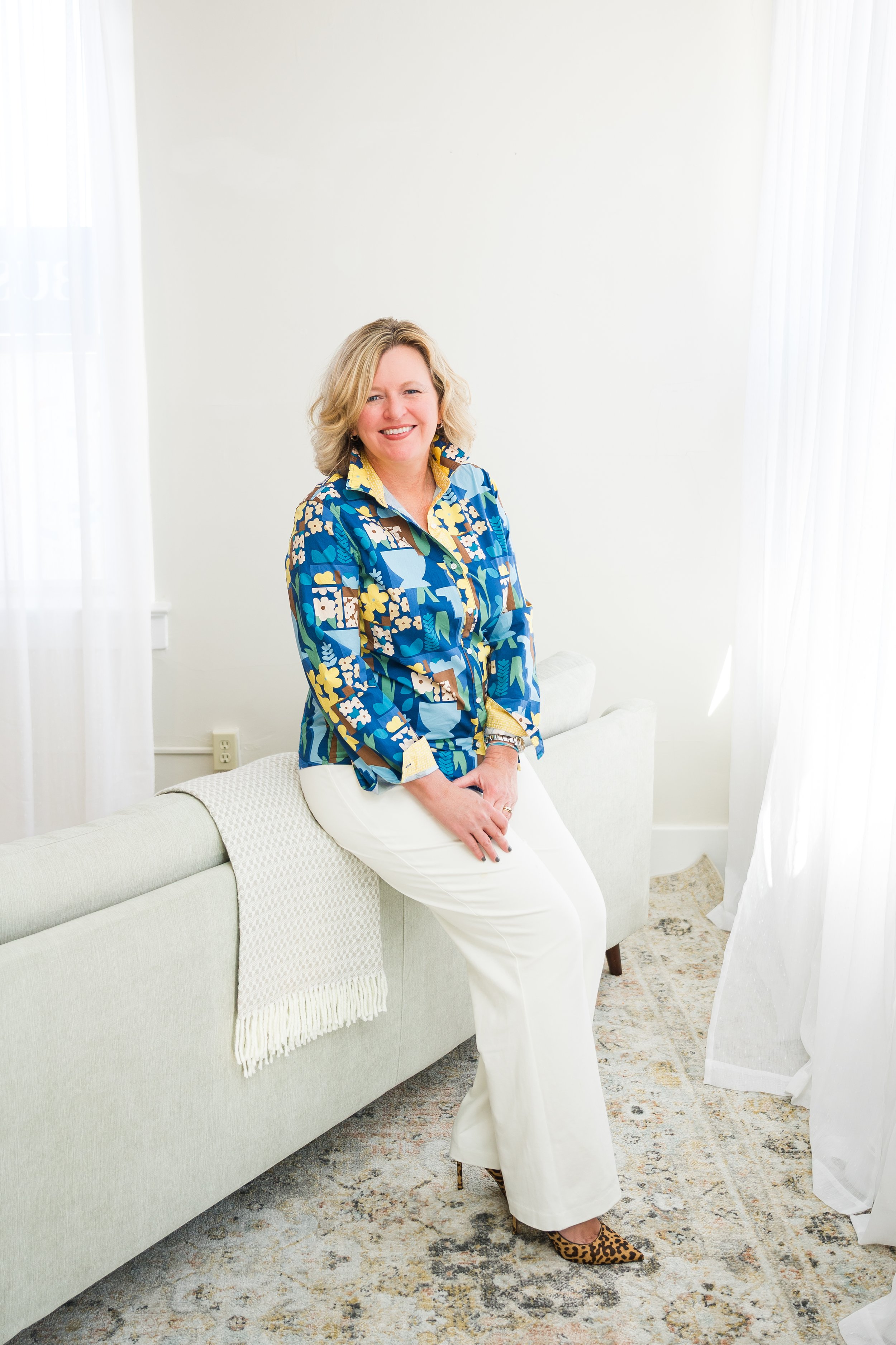 A woman with blonde hair wearing a colorful blue, yellow, and white patterned blouse and white pants, sitting on the arm of a light-colored sofa in a bright room with white curtains.