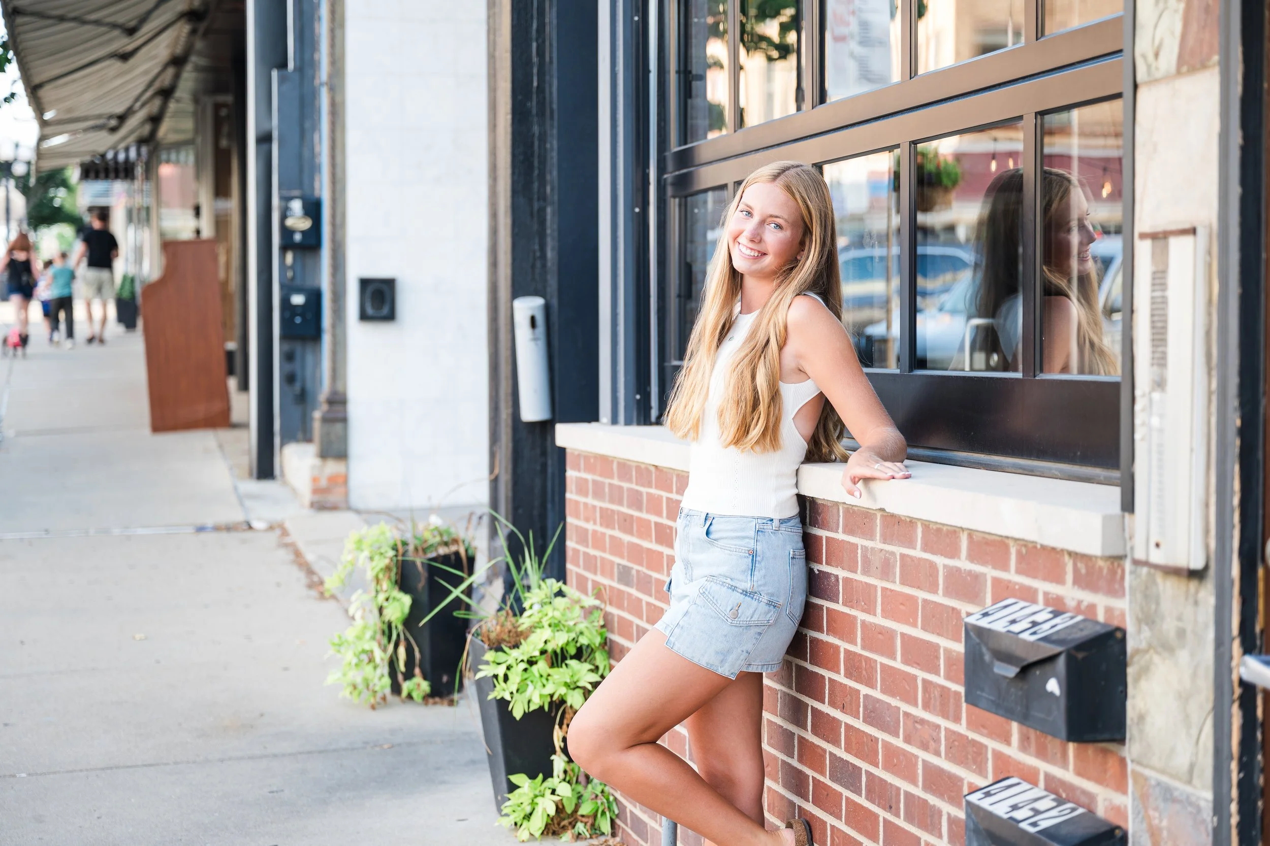 A young woman with long blonde hair wearing a white sleeveless top and denim shorts, leaning against a brick wall outside a building, smiling at the camera.