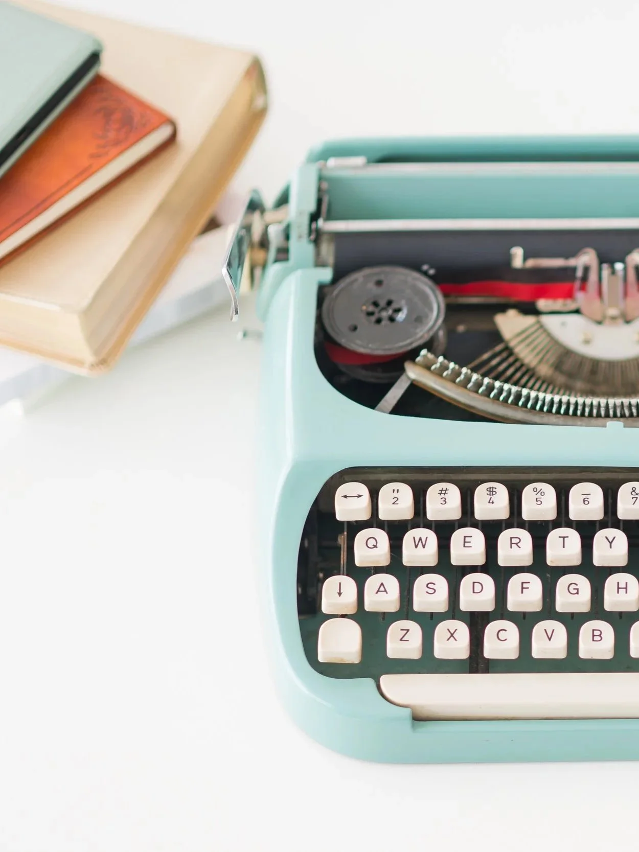 Bloomington Normal IL business photographer, A vintage turquoise typewriter with white keys, placed on a white surface, with a stack of books nearby.