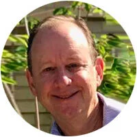 A smiling man with short hair and a beard, outdoors with greenery in the background.