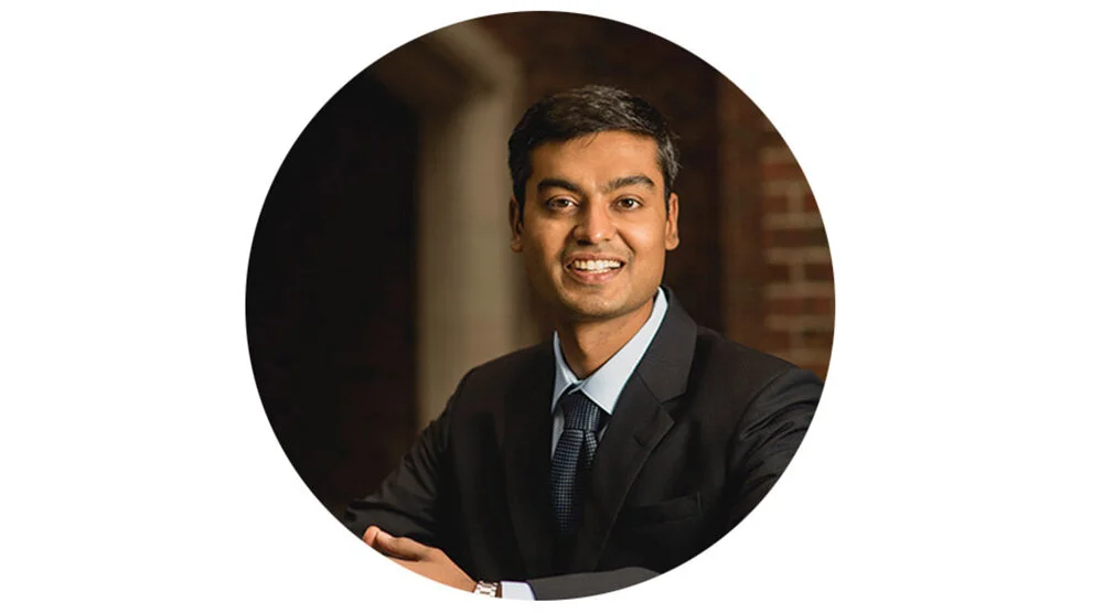 A man with dark hair in a suit smiling with arms crossed, sitting indoors with a brick wall in the background.