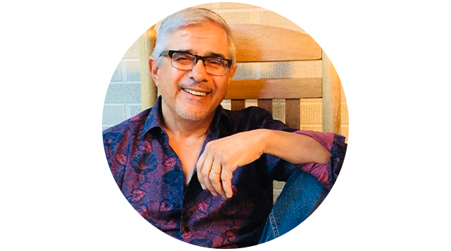 A smiling man with glasses and gray hair sitting in a wooden chair, wearing a patterned shirt, in an indoor setting.