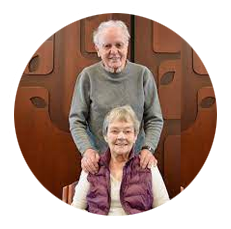 An elderly man and woman smiling, with the man standing behind the woman who is seated.
