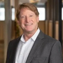 A smiling man with light brown hair, wearing a gray suit jacket and a checkered shirt, standing indoors.