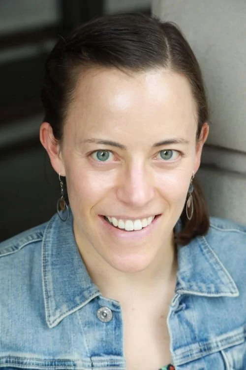 Close-up of a woman with short brown hair, blue eyes, and earrings, smiling, wearing a denim jacket.