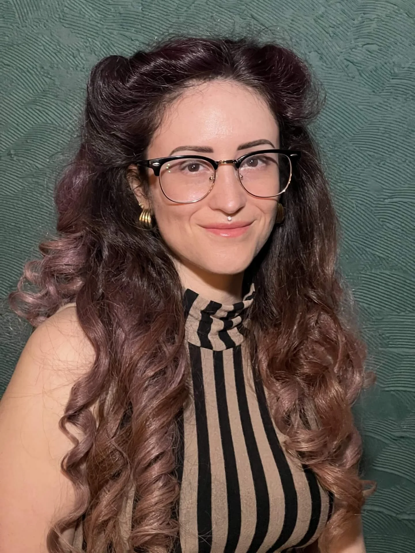 A woman with long, curly hair with pinkish-brown highlights, wearing glasses, gold hoop earrings, and a black and beige striped sleeveless top standing against a textured green wall.