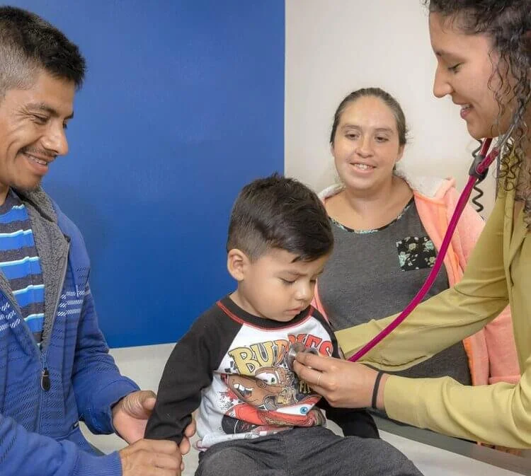 A young boy visits a doctor with his parents. The doctor, wearing a yellow coat, uses a stethoscope to check the boy's heartbeat. The boy is sitting calmly, and his parents are smiling and supporting him.