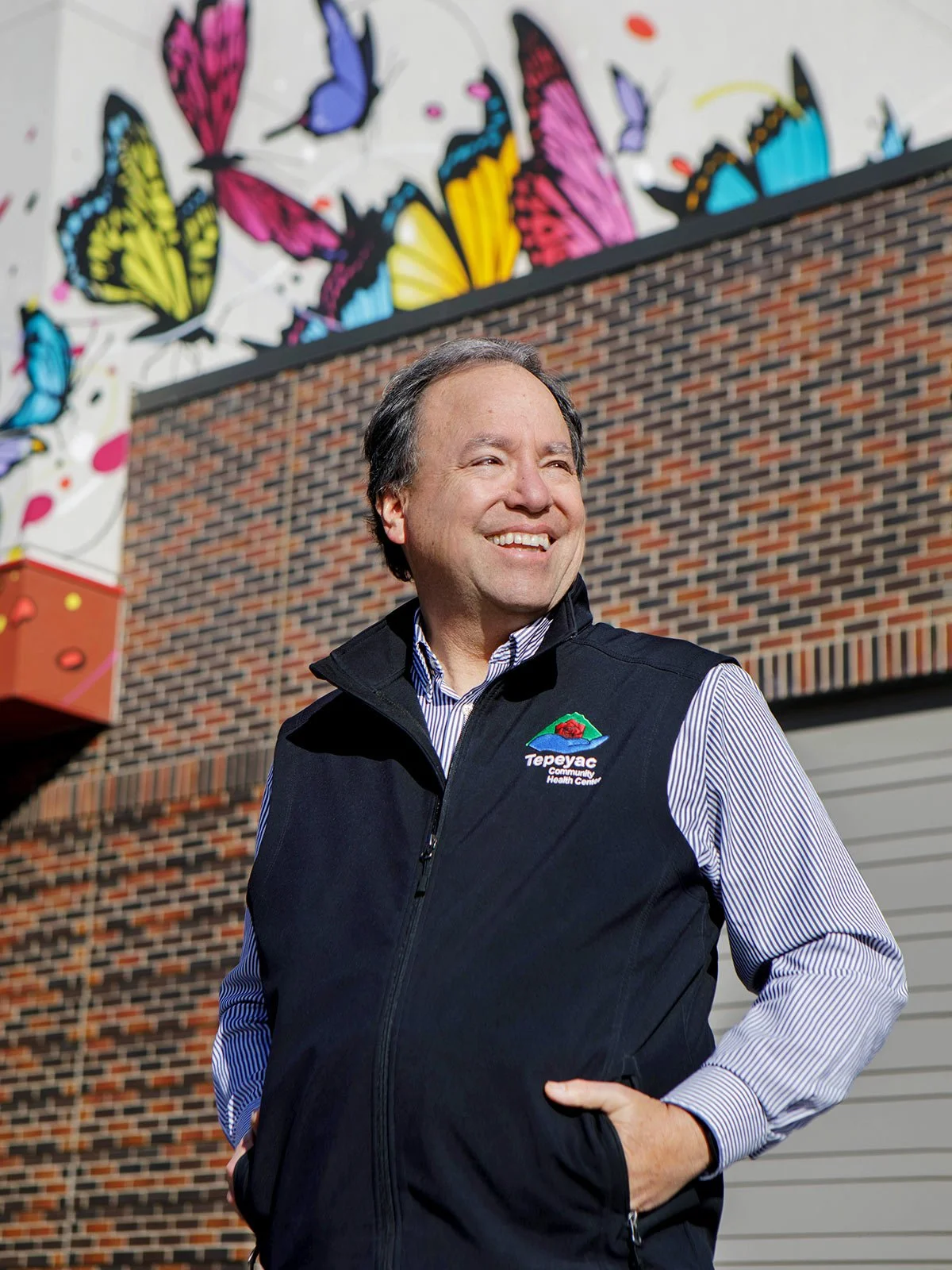 A smiling man stands outdoors in front of a brick wall with colorful butterfly artwork.