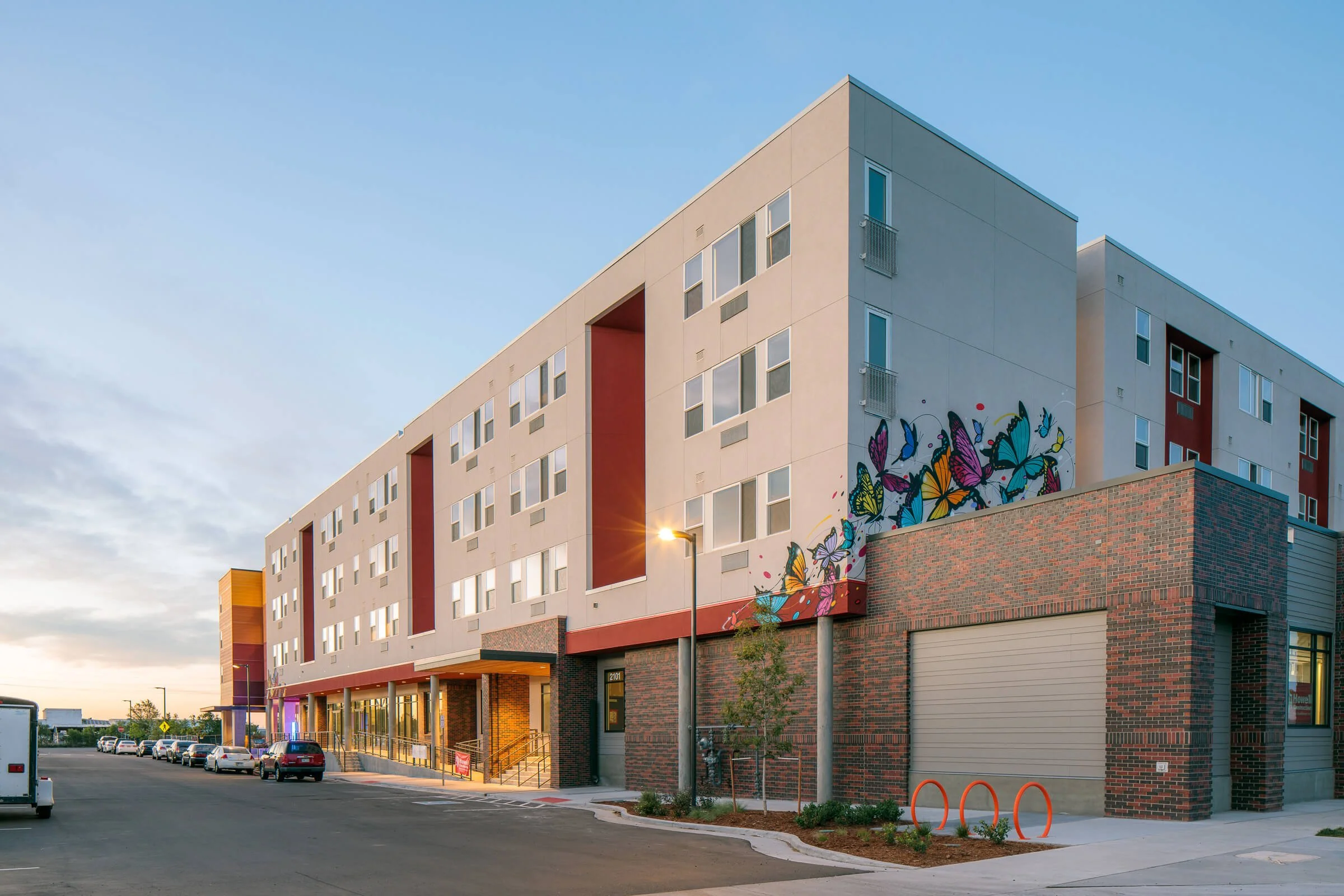 The complex where Tepeyac is located off 48th and Vine Street. There is a mural of colorful butterflies on its white exterior wall, featuring a ground-level health center, a bus stop, and a parking lot with several cars and pedestrians.