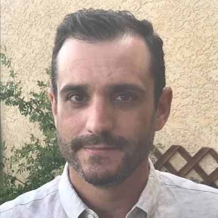 A man with dark hair and a beard, wearing a light-colored button-up shirt, standing outdoors with a beige wall and greenery in the background.