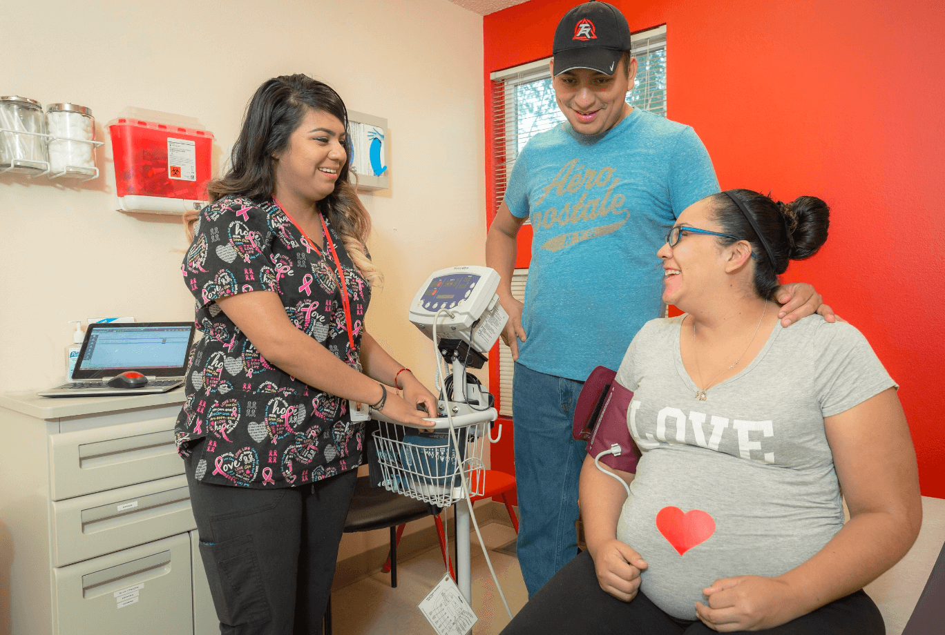 A nurse takes a pregnant woman's blood pressure while a man stands nearby, smiling, in a medical clinic. The woman is seated, wearing glasses and a gray shirt with a red heart, and the man has his hand on her shoulder.