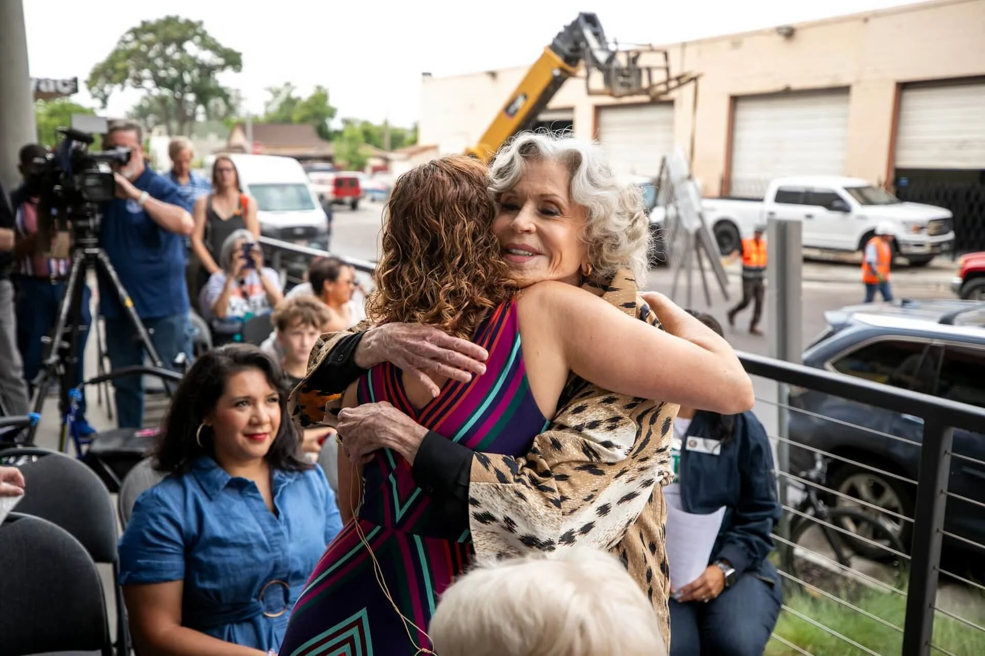 Actress and activist Jane Fonda hugs a women at a rally gathering outdoors, with several people, vehicles, and a construction vehicle in the background.