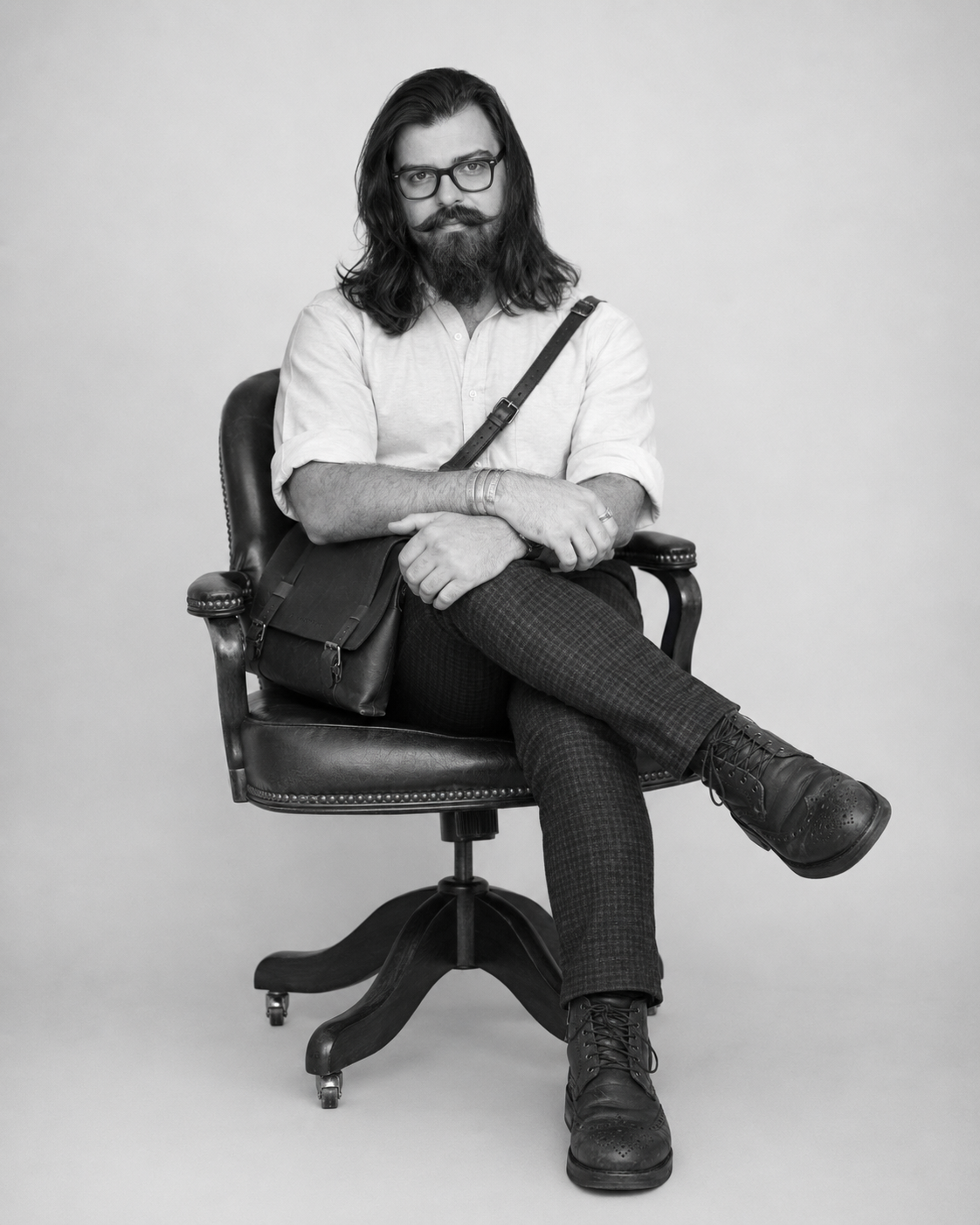 Black-and-white photo of a man with long hair and glasses sitting in an office chair. He has a beard and mustache, is wearing a light-colored shirt, patterned pants, and black shoes. He has a bag on his lap and is posing with his legs crossed.