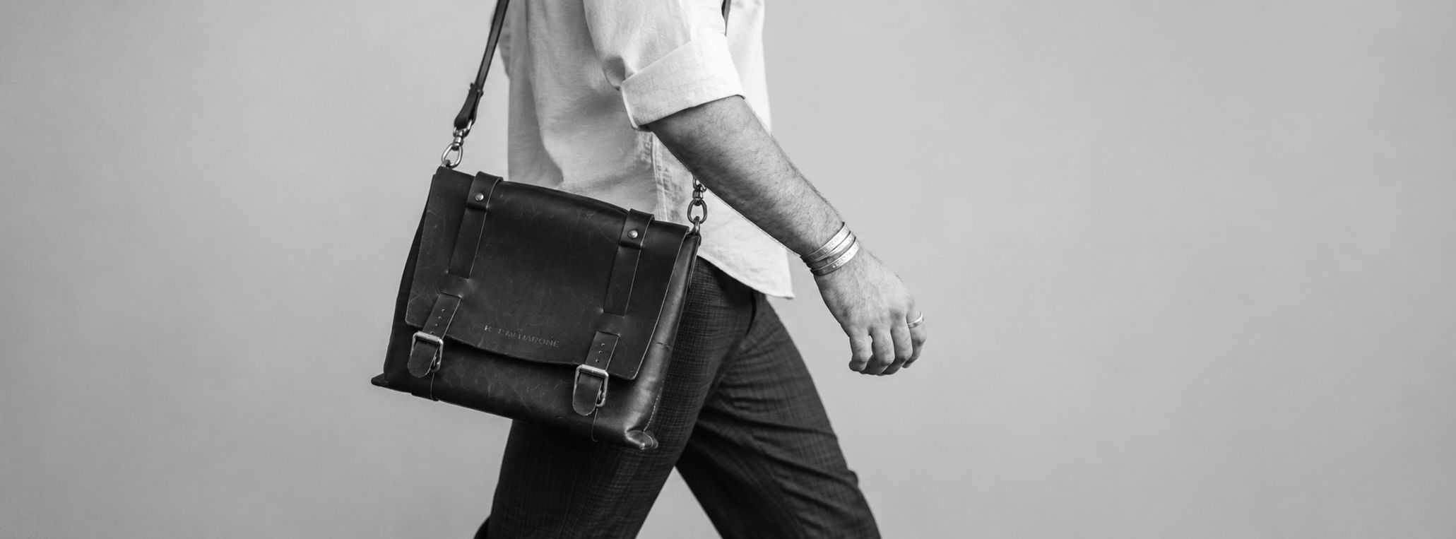 A man walking with a black leather bag over his shoulder, wearing a light-colored shirt with rolled-up sleeves and dark plaid pants, against a plain light background.