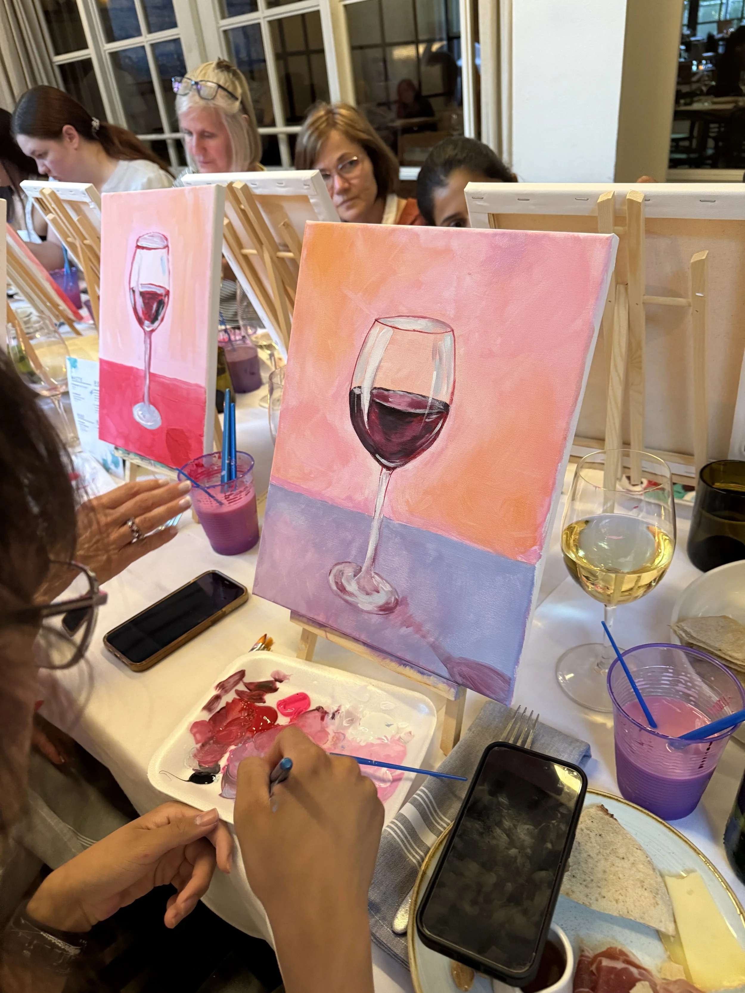 People participating in a painting class, painting wine glasses on small canvases, with paints, brushes, and drinks on the table.