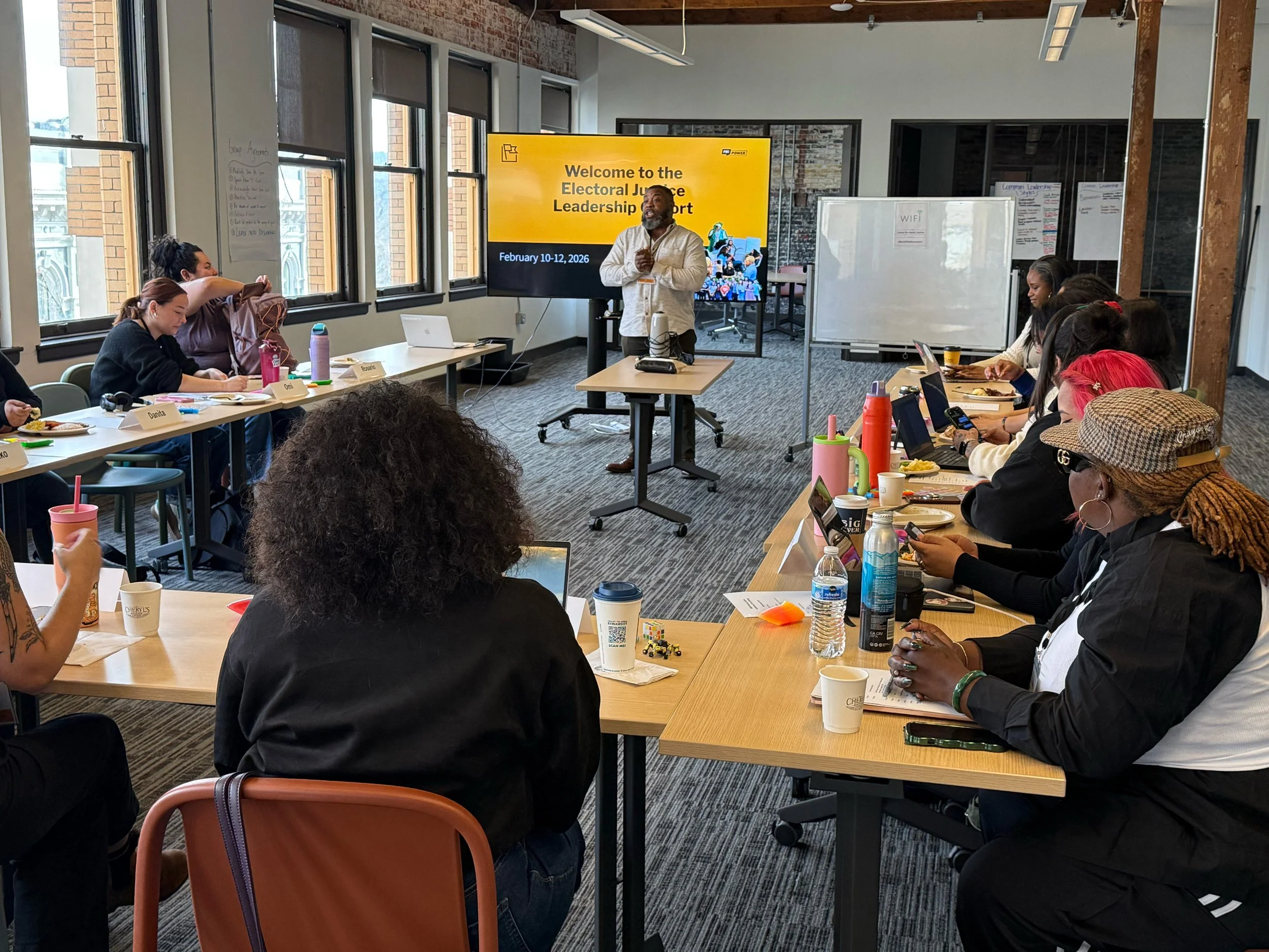 A dozen people sit at tables arranged in a U shape. A person stands and speaks at the front of the U in front of a television screen with the words "Welcome to the Electoral Justice Leadership Cohort" projected onto it.