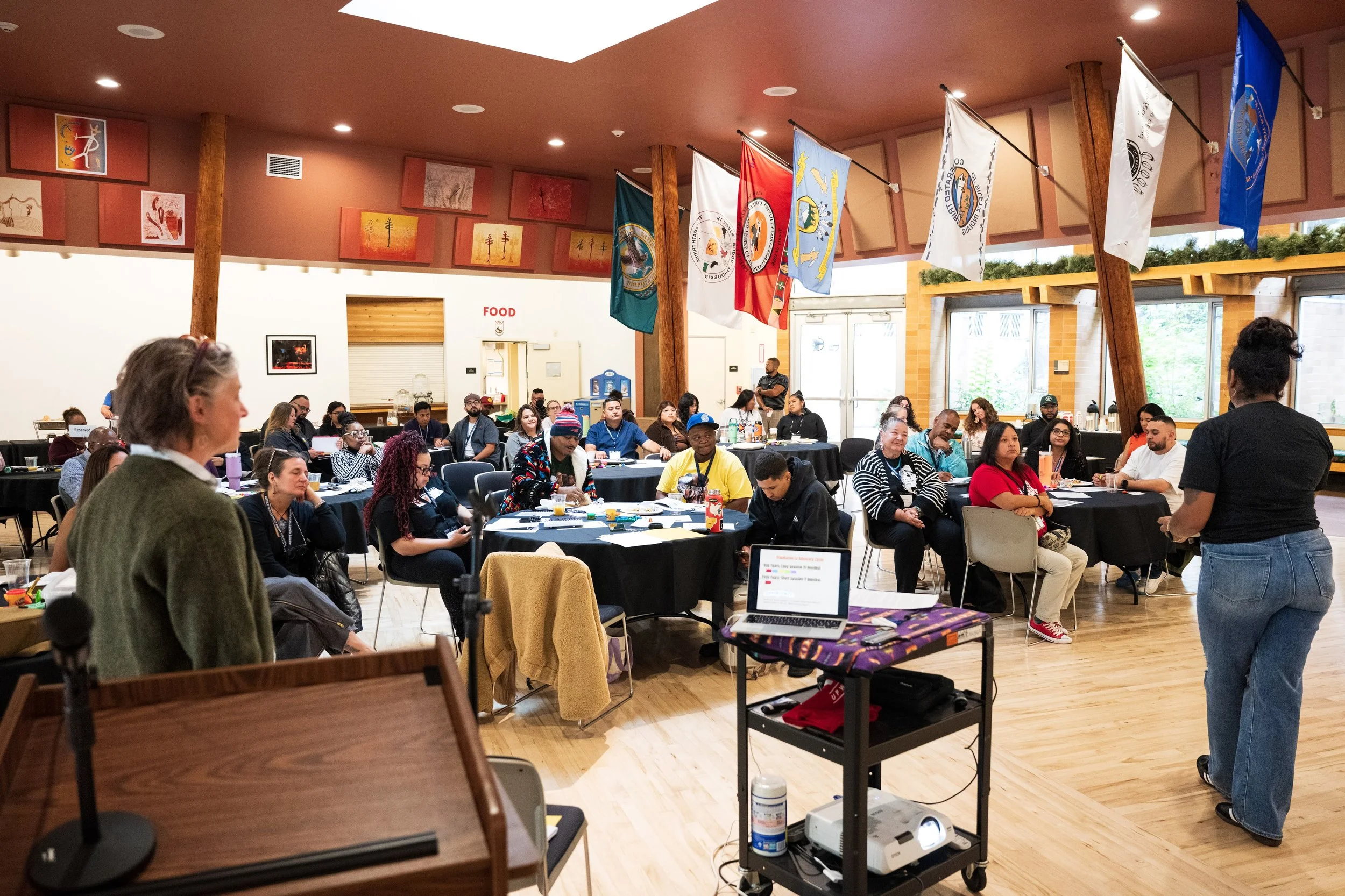People sit at round tables inside a room decorated with Native art and tribal flags.