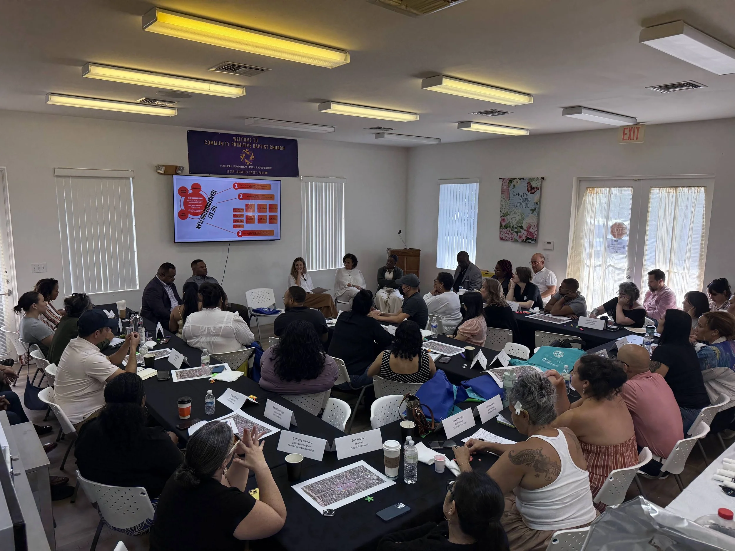 A couple dozen people packed into a room seated around tables, all watching a screen and a panel of four people seated underneath the screen speaking into microphones.
