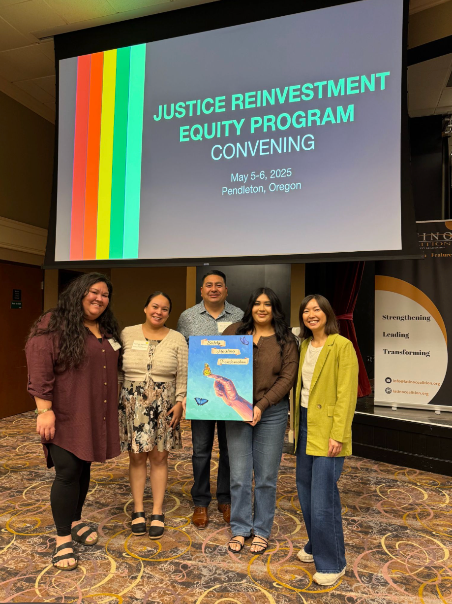 Cleo Tung and JREP participants pose for a photo holding a poster with butterflies held in a hand and pose in front of a slideshow presentation titled Justice Reinvestment Equity Program Convening May 5-6 2025, Portland Oregon