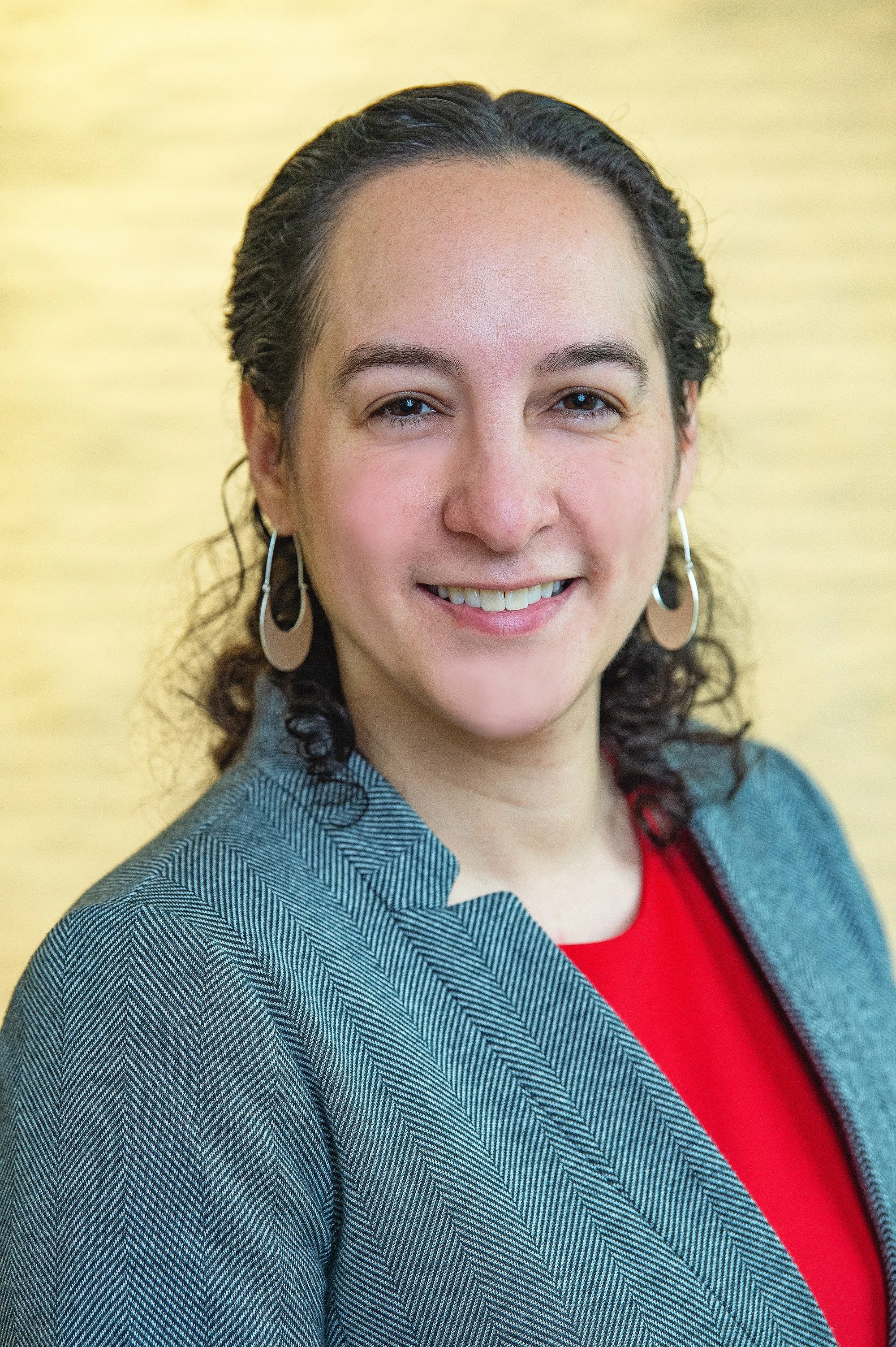 Professional photo portrait of a light-skinned Latina woman with dark curly hair wearing a tweed blazer and smiling.