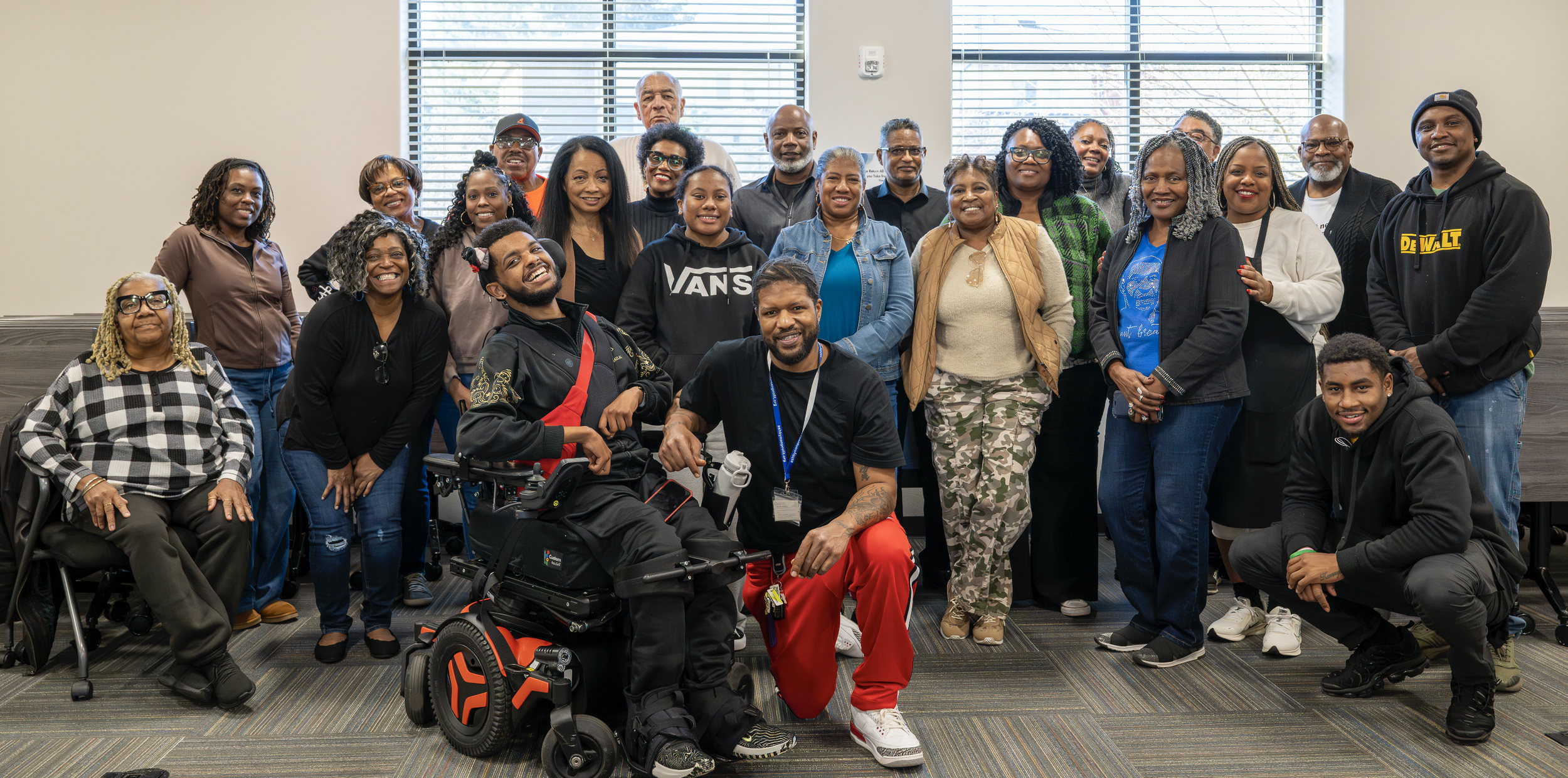 Photo provided by Ashby Family Lead & Engagement: a Black man in a motorized wheelchair poses with Black man in red pants surrounded by smiling community members and staff
