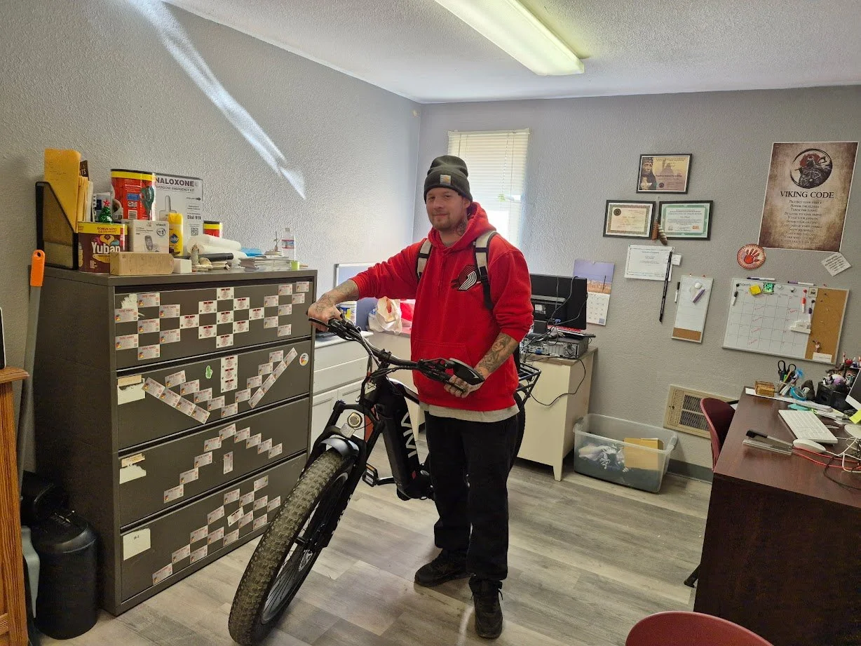 A man wearing a Portland Trailblazers hoodie stands in the middle of an office holding the handle bars of an e-bike.