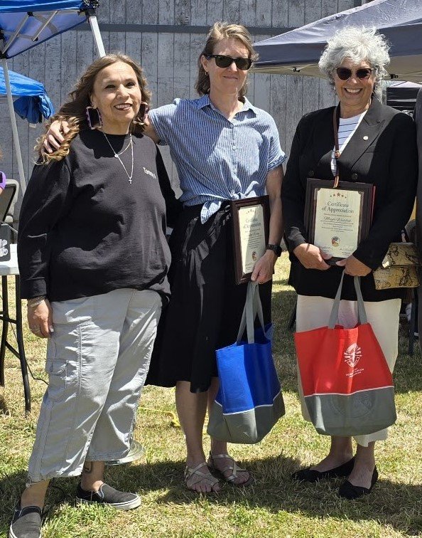 Three women stand together smiling. Two hold tote bags and certificates of appreciation.