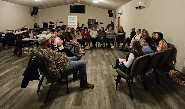 A couple dozen people sit in a circle of chairs.