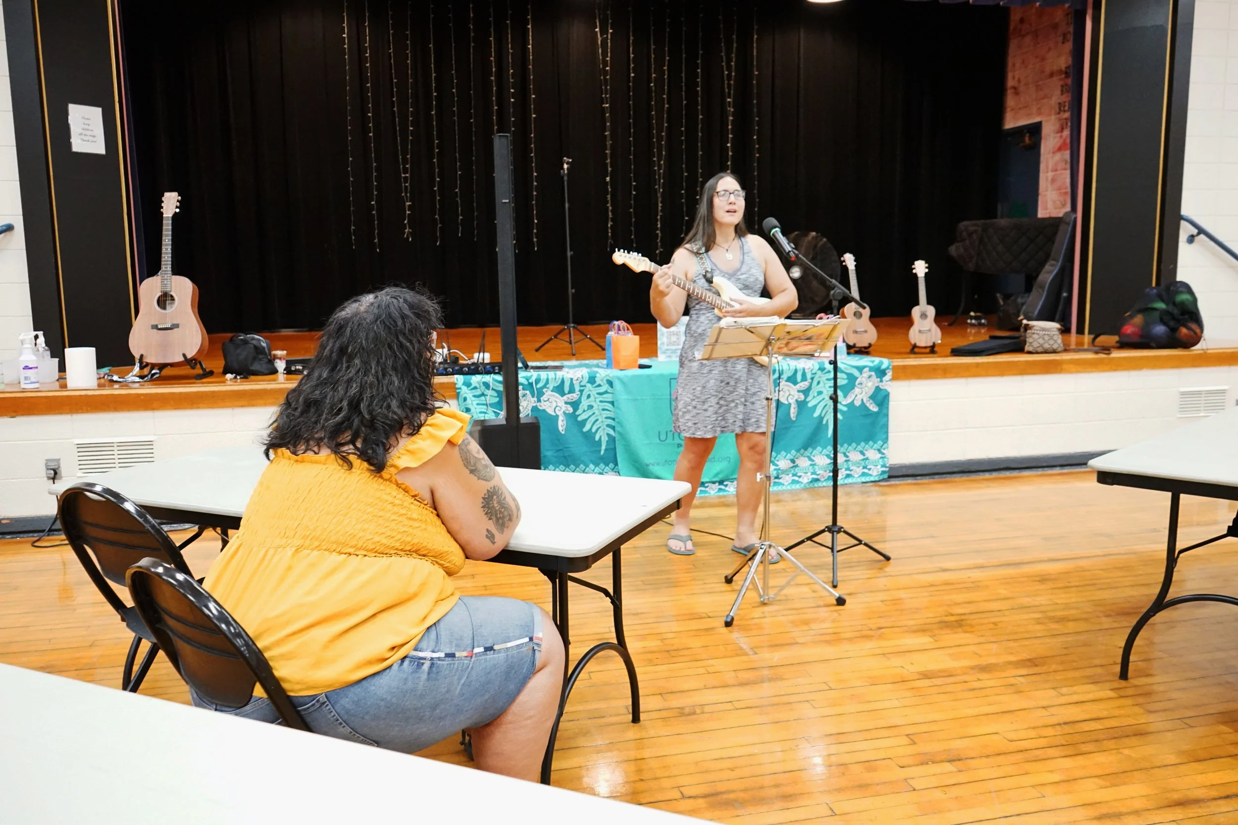 A person seated at a folding table leans forward as they listen to a person standing in front of a stage playing a guitar.
