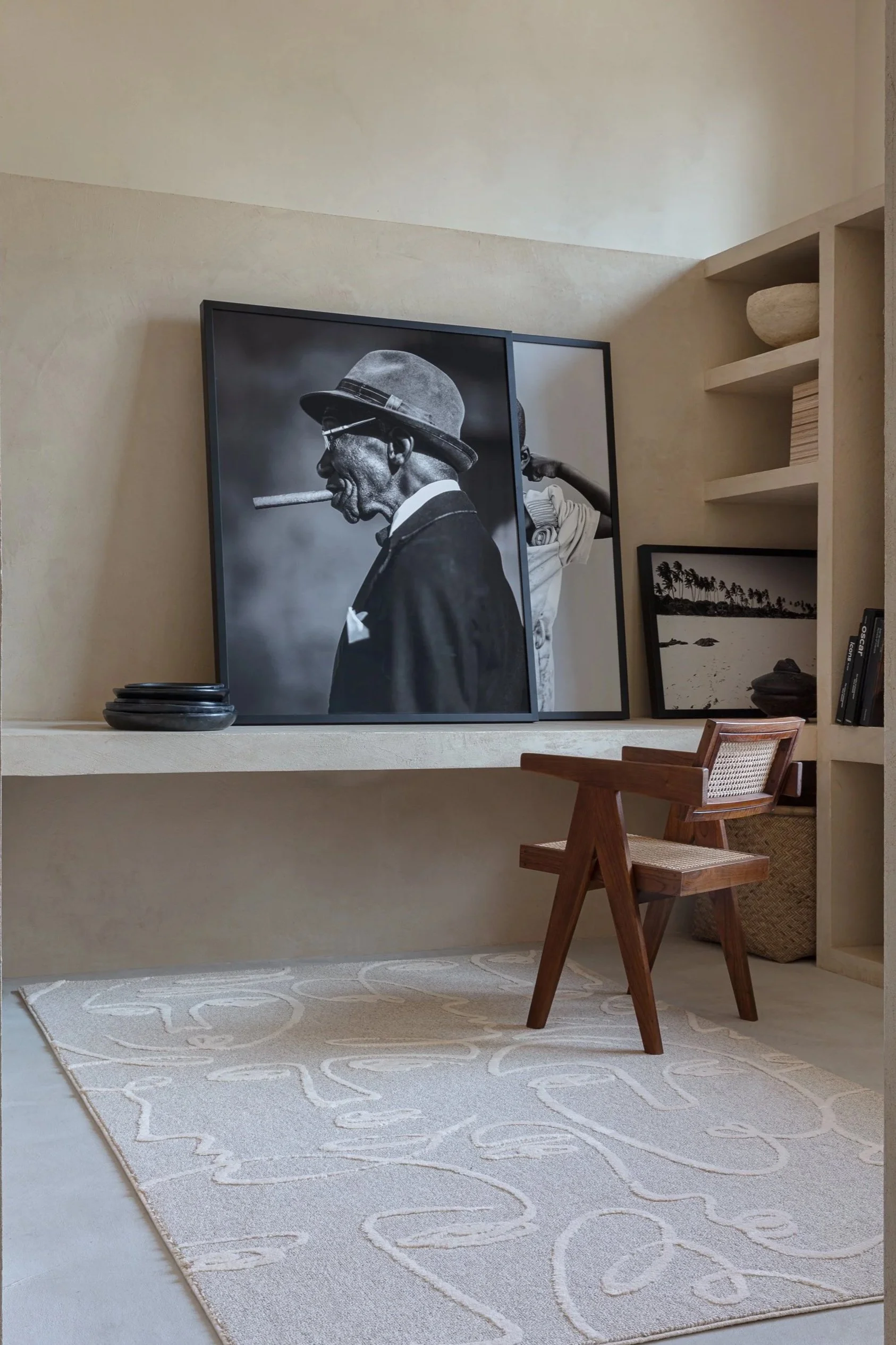 Interior room with a beige wall, featuring a large black and white framed portrait of a man in a suit and fedora smoking a cigar, a short wooden chair, a white patterned rug, and built-in shelving with various decorative items.