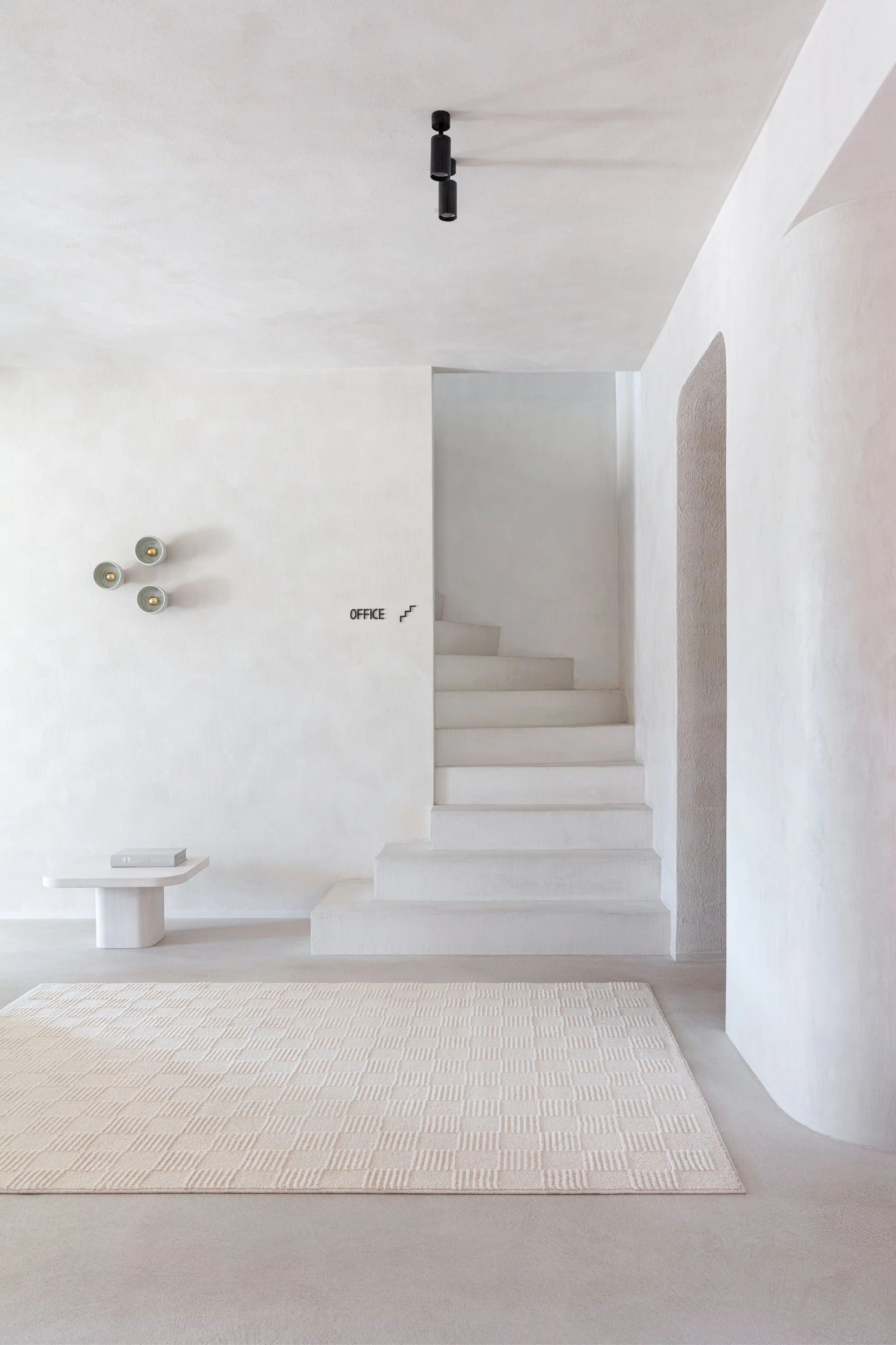 Minimalist white interior with staircase leading to an upstairs office, beige rug on the floor, small white table with a book, and modern wall lamps.