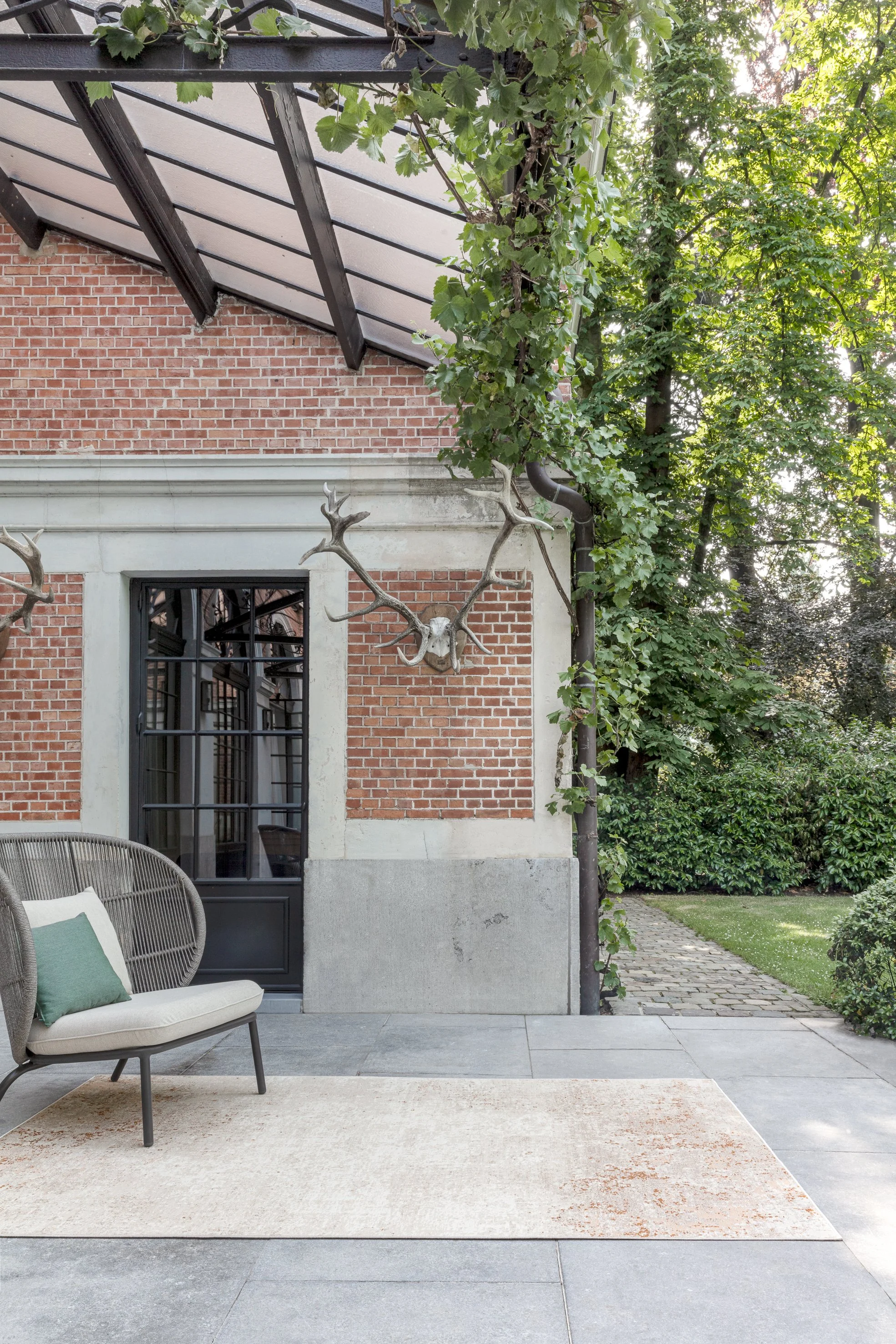 Outdoor patio area with a modern chair and a decorative antler wall mount on a brick and concrete wall, surrounded by green trees and plants.