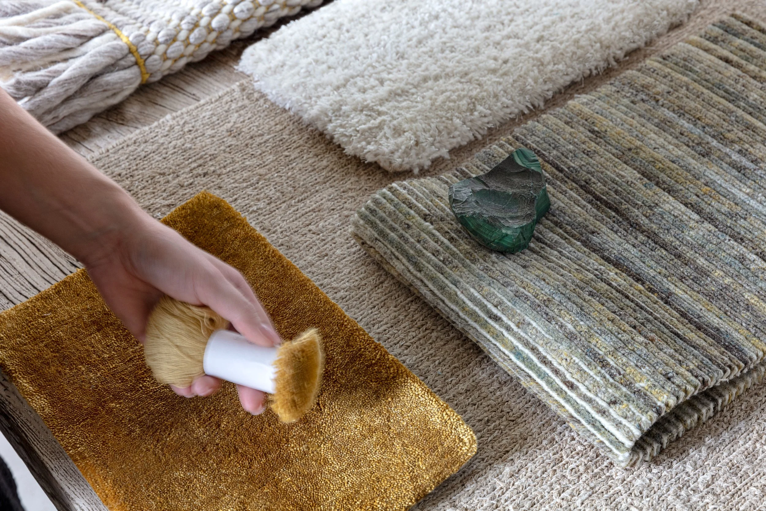 Person using a soft brush to clean a yellow-textured rug on a table with various rugs and a green stone.