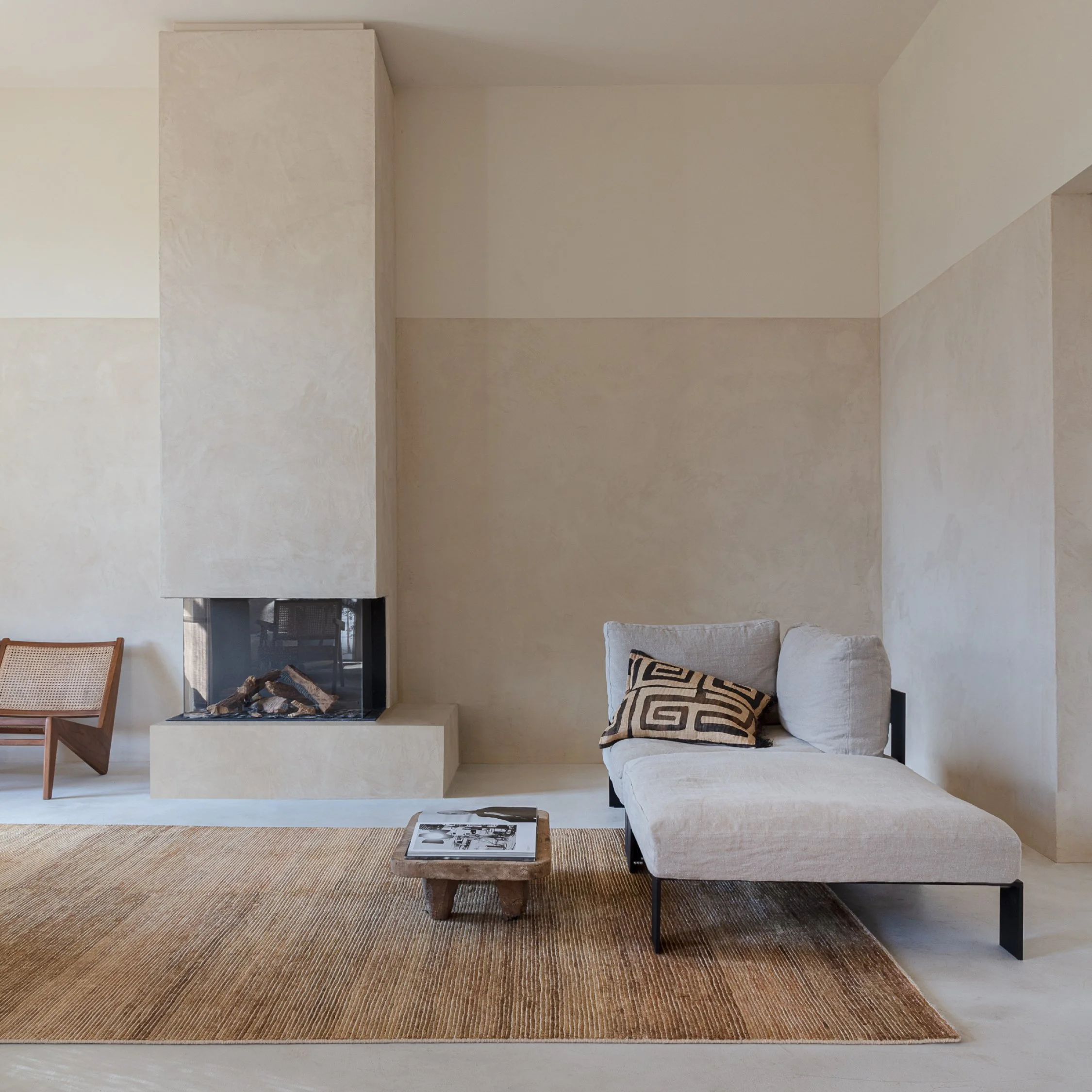 Minimalist living room with a fireplace, a chaise lounge with pillows, a wooden side table with a magazine, and a woven rug.