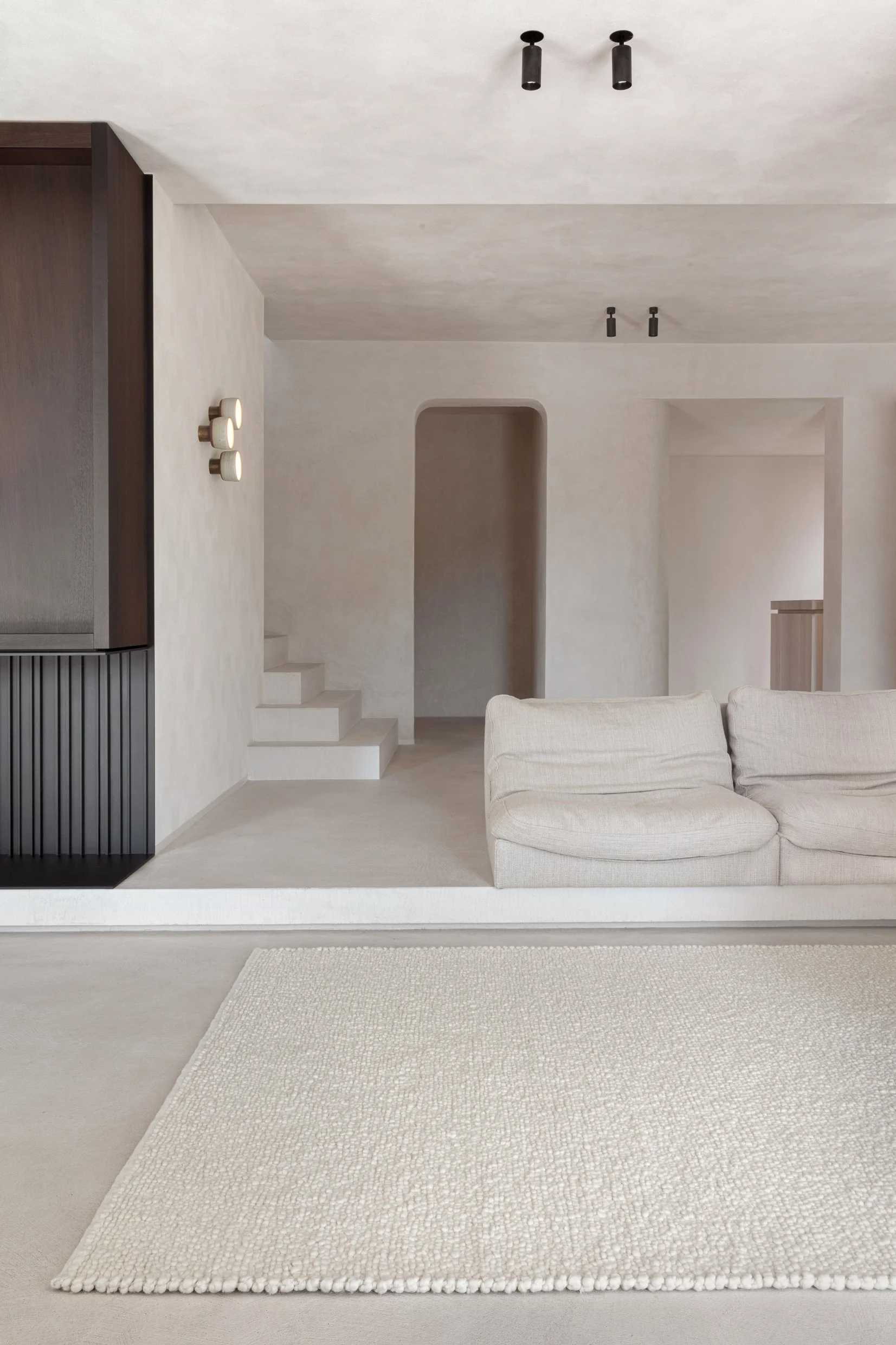 A minimalist living room features a beige sofa, a textured white area rug, and neutral-toned walls with modern lighting fixtures.