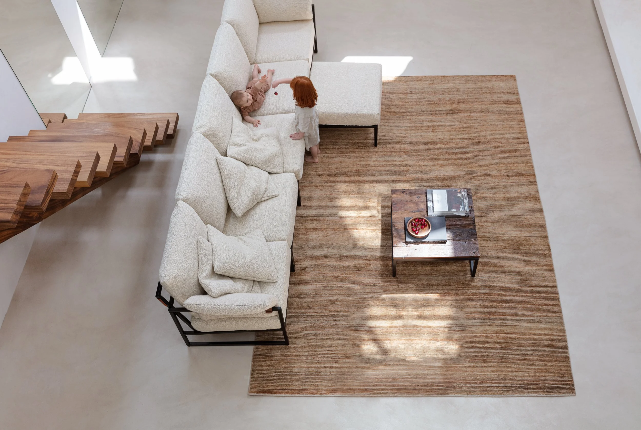A top-down view of a modern living room with a beige sectional sofa, a brown area rug, a wooden coffee table with a cake and magazines, and two small children playing on the sofa near a staircase.