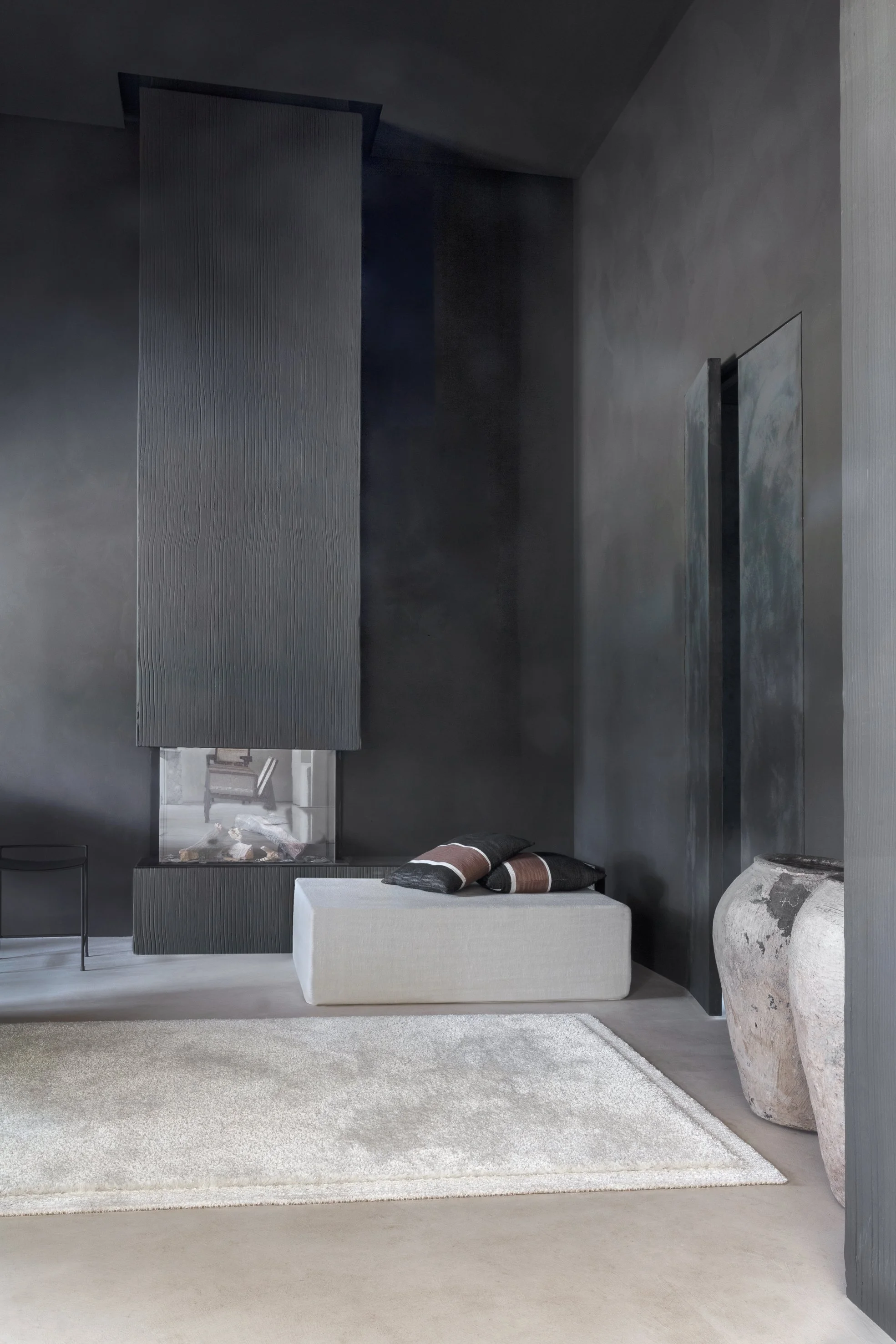 Minimalist interior with dark walls, a light-colored bench with pillows, a white rug, and large ceramic vases.