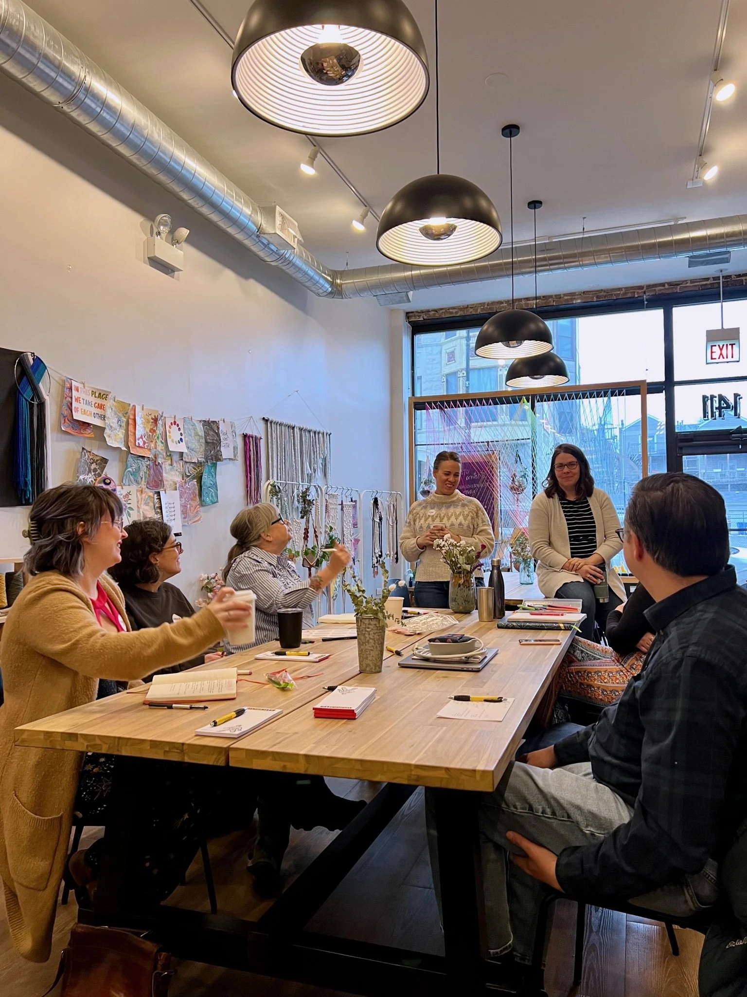 a group of men and women business owners sit around a wooden table surrounded by colorful artwork in a bright, airy studio workspace.