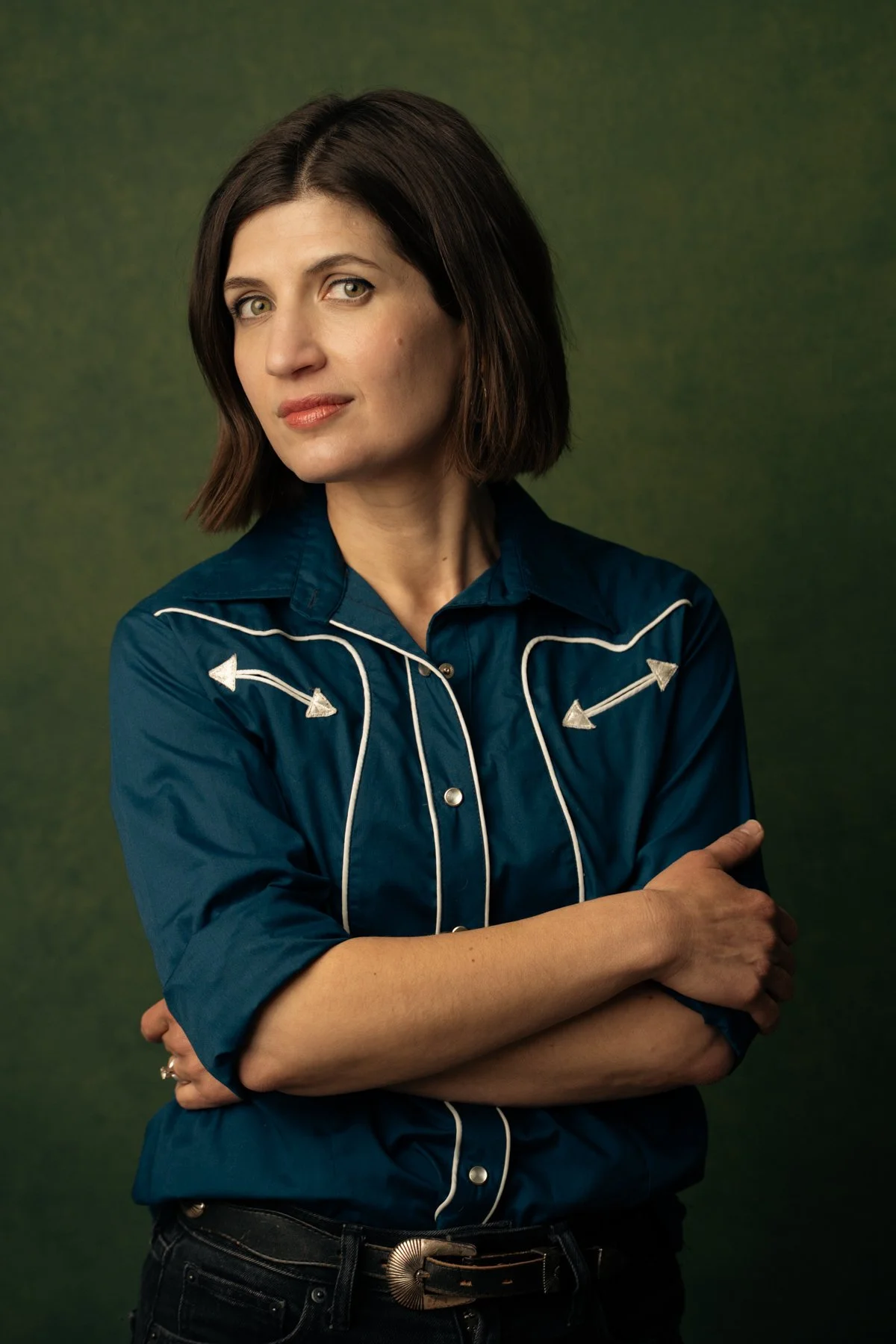 A woman with short brown hair and light skin standing against a textured green background, wearing a vintage blue Western-style shirt with white piping and arrow embroidery, and a black belt with a large decorative buckle.